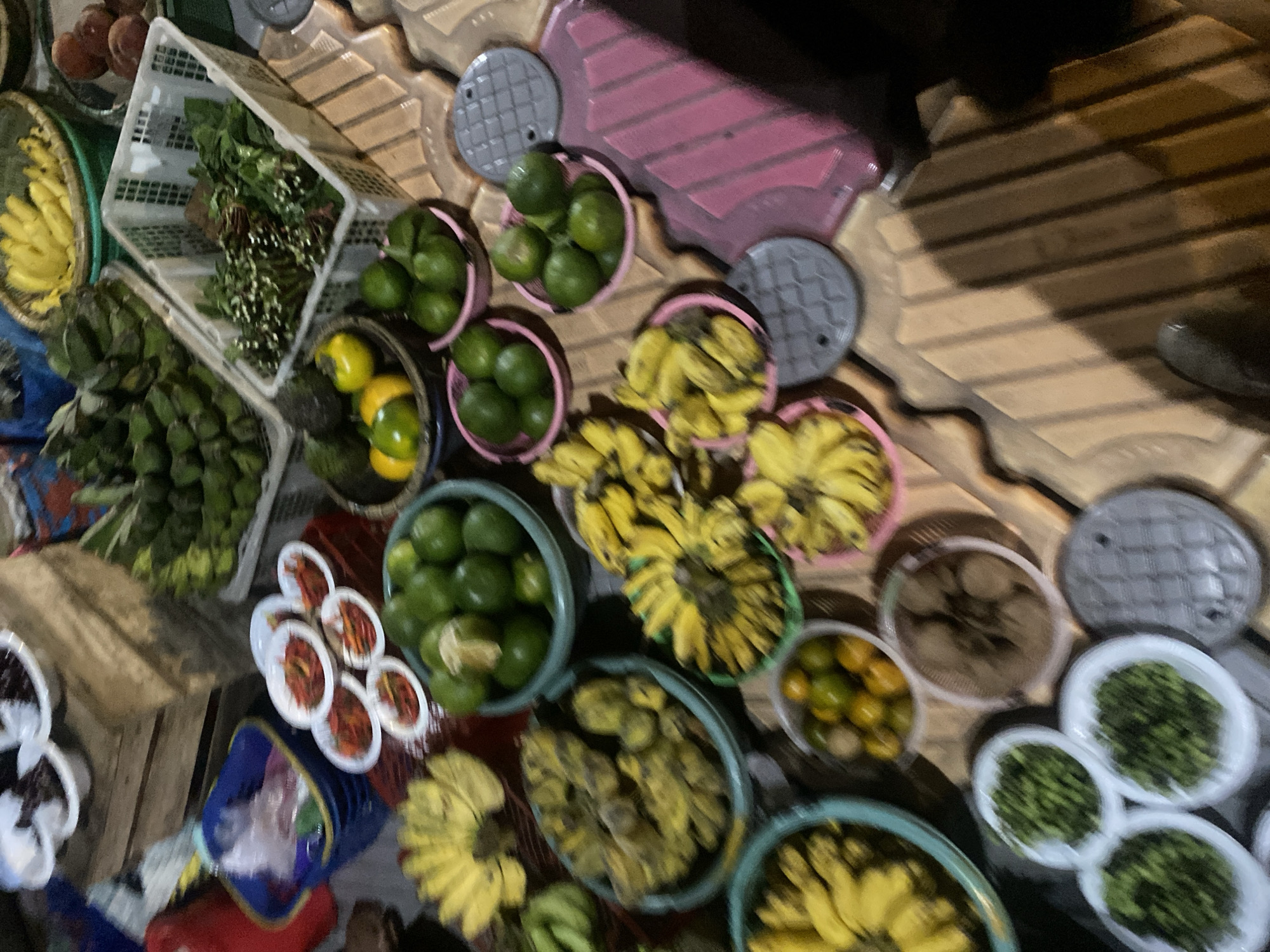 The image depicts a night market scene in Banjarmasin, Indonesia, showcasing a variety of fresh produce for sale. In the foreground, several bowls and baskets overflow with colorful fruits and vegetables. Prominently displayed are bunches of ripe yellow bananas, some appearing overripe, and green limes arranged in blue and pink containers. Small, round citrus fruits, possibly tangerines or kumquats, are gathered in a white bowl, alongside dried goods in a woven basket. Further back, a selection of red chilies is meticulously arranged in small white dishes. The market stall also features green leafy vegetables and other unidentifiable produce, some in white plastic crates and woven baskets. The setting is dimly lit, typical of an outdoor night market, with shadows cast across the patterned surfaces of the stall and surrounding ground. The overall impression is one of a bustling marketplace offering a diverse array of fresh goods.