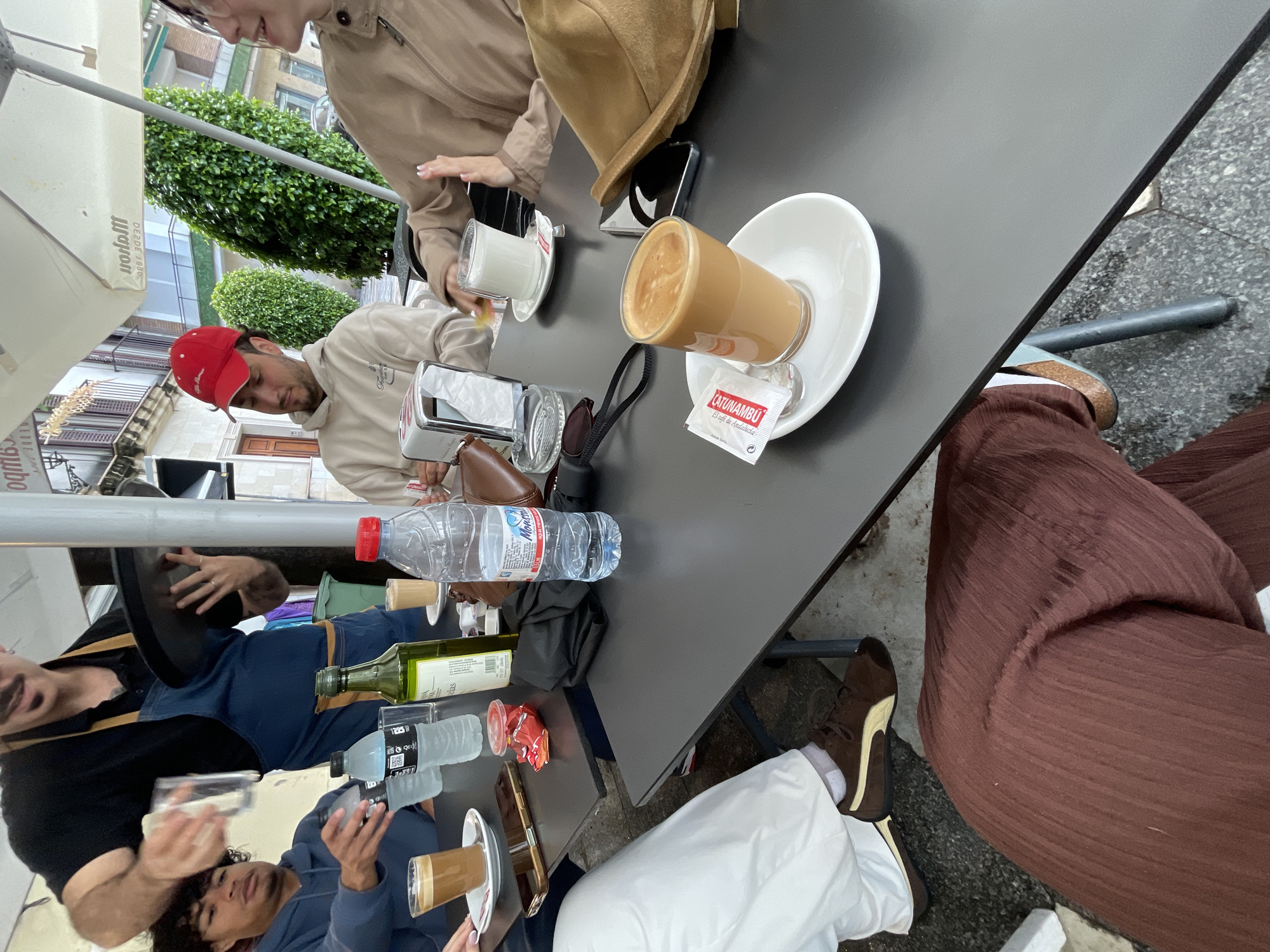 The image captures a street-side cafe scene in Jerez De La Frontera, Spain. The shot is taken from an elevated angle, looking down at a dark gray table top. Several individuals are seated around the table, their bodies partially visible. A man wears a red baseball cap and a light-colored sweatshirt. Another person is wearing a tan jacket and a brown hat. Another person is wearing brown pants and brown and white shoes. A waiter, dressed in a black shirt and blue jeans, serves customers, holding a tray in his left hand and carrying a glass in his right hand. A second waiter wears a blue jacket and is holding a glass.
Various items are placed on the table, including glasses of coffee, water bottles, a bottle of olive oil, sugar packets, a wallet, and cellphones. The cafe is located outdoors, as suggested by the presence of an umbrella. A building facade and lush greenery can be observed in the background, indicative of a city street.