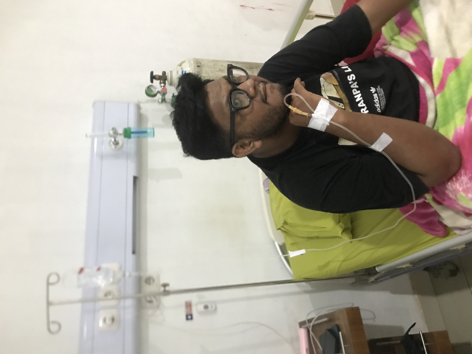 A man wearing glasses and a black t-shirt with the Adidas logo lies in a hospital bed, an IV drip inserted into his arm. The room is sterile and appears to be in a hospital setting. To the left, hospital equipment is visible, including an oxygen tank and a fluid stand. The patient is covered with a colorful blanket, and the walls are white. Considering the prompt, the location is implied to be in Lhokseumawe, Indonesia.