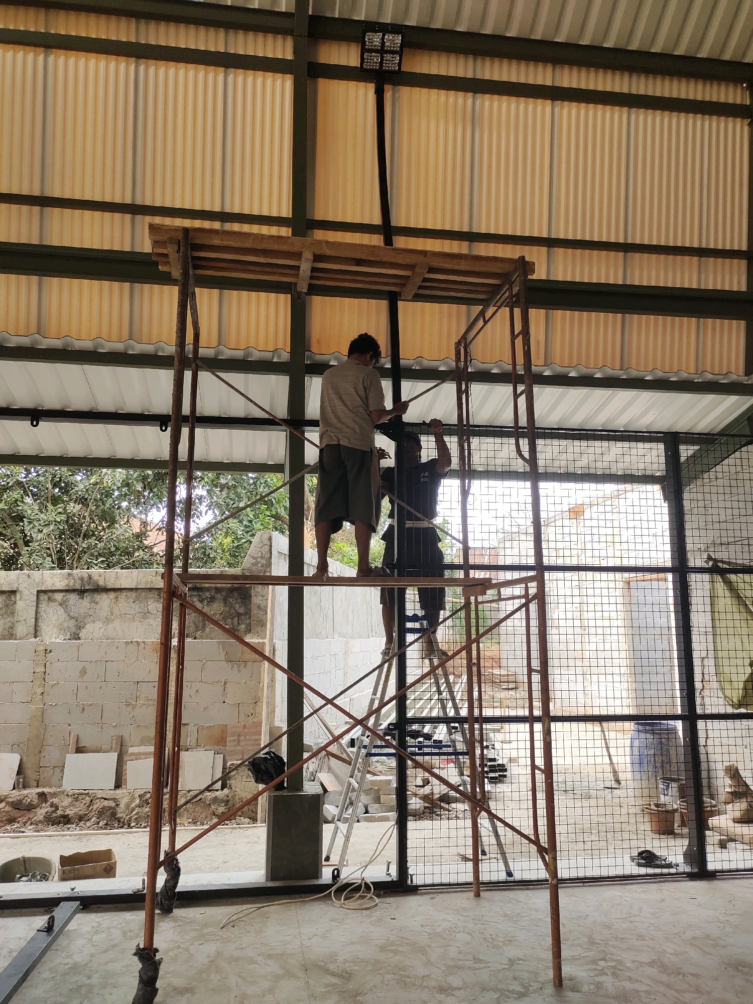 Two individuals are working on scaffolding within a partially constructed building. The structure has a corrugated metal roof and appears to be an open-air or semi-enclosed space, with a wire mesh partition in the background. One person, wearing a light-colored shirt and shorts, stands on a higher platform of the scaffolding, holding a piece of wood. Another person, in a dark t-shirt and shorts, is positioned below and slightly to the side on the scaffolding, appearing to be assisting. In the background, there are building materials, including concrete blocks, and some vegetation is visible through the open areas. The scene suggests ongoing construction or renovation work at a site in Lengkong Kulon, Indonesia.