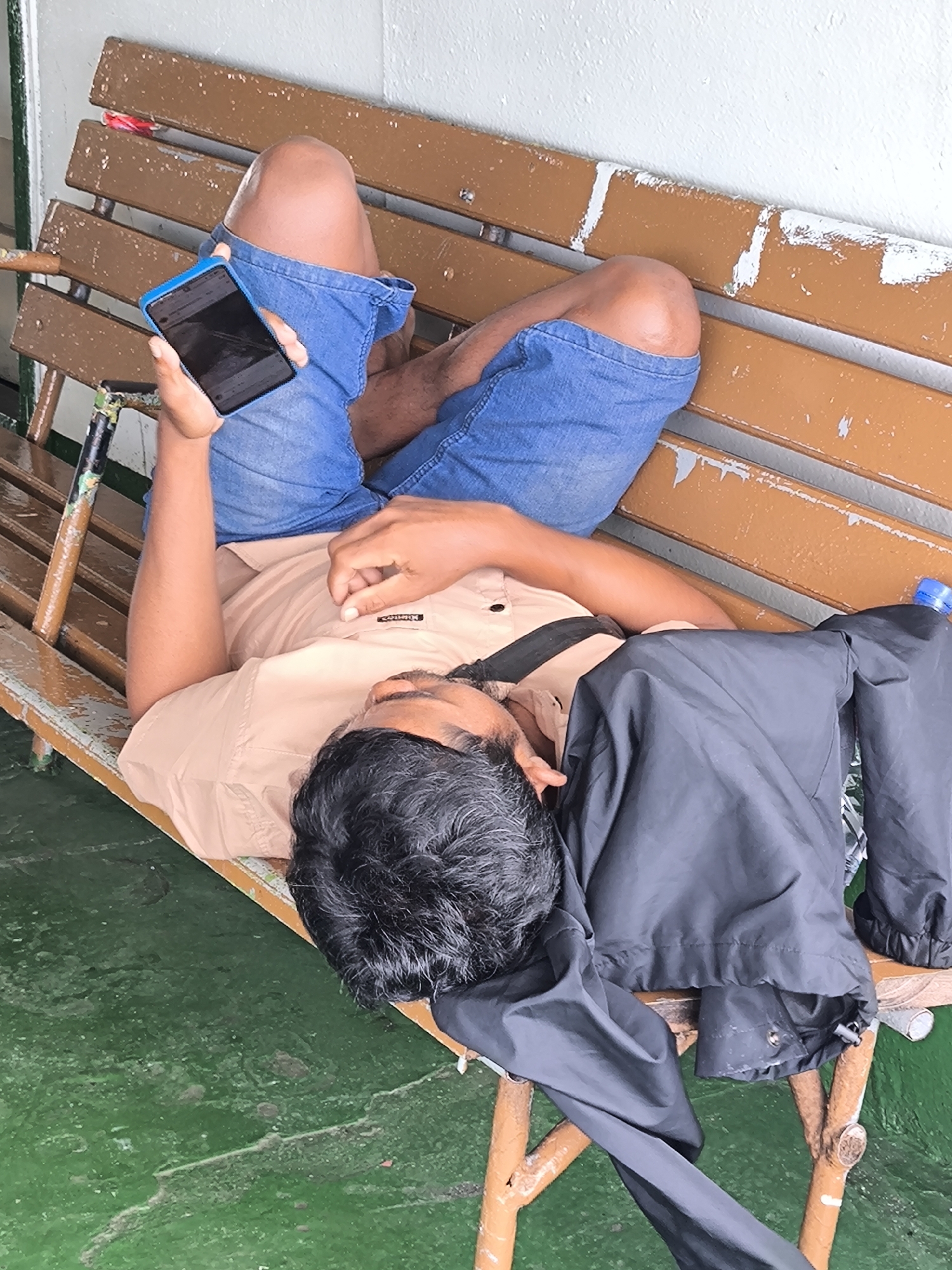 A man lies on a wooden bench while holding a blue-cased smartphone in his right hand. The bench has a worn brown paint job, and the man's legs are crossed on it, wearing blue denim shorts. He is wearing a tan short-sleeved shirt, and a black strap is visible across his chest. A folded black jacket is draped to his side. The floor beneath the bench is painted green. The location may be in the city of Lembar Selatan, Indonesia.