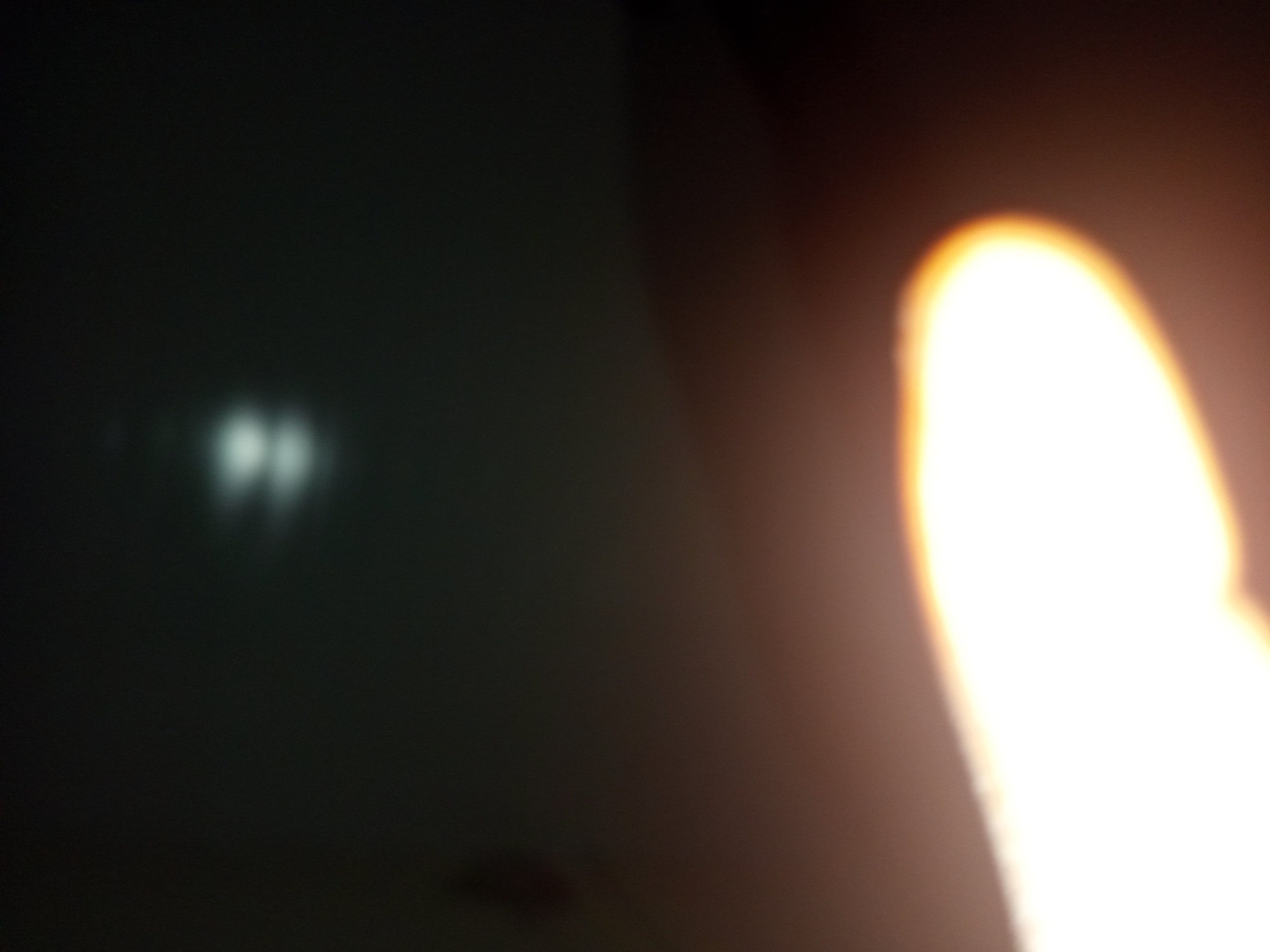 This is a low-light, blurred photograph that appears to be taken indoors. In the upper portion of the image, two bright, indistinct white lights are visible against a dark background. In the lower portion of the image, a large, brightly illuminated area, possibly a light source or a reflection, dominates the frame. There is a gradient of light to dark from the bottom left to the top right. There are no recognizable landmarks or contextual clues to identify the specific location within Jalingo, Nigeria. The image quality is too poor to discern any people, objects, or actions.