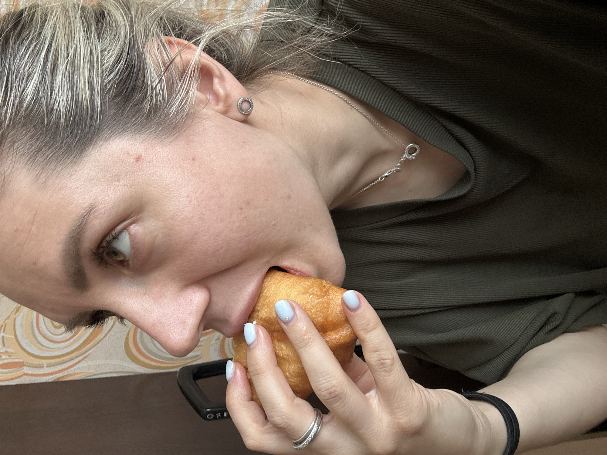 The image is a close-up, side-profile shot of a Caucasian woman with blonde hair, taking a bite out of a fried pastry. She is wearing a dark green, ribbed t-shirt, a silver ring on her left hand, and a silver necklace with a circular pendant. Her fingernails are painted a light blue. In the background, a wall with a retro-style patterned wallpaper is visible. The lighting suggests an indoor setting. There is no identifiable context to suggest the location is Novoaltaysk, Russia.