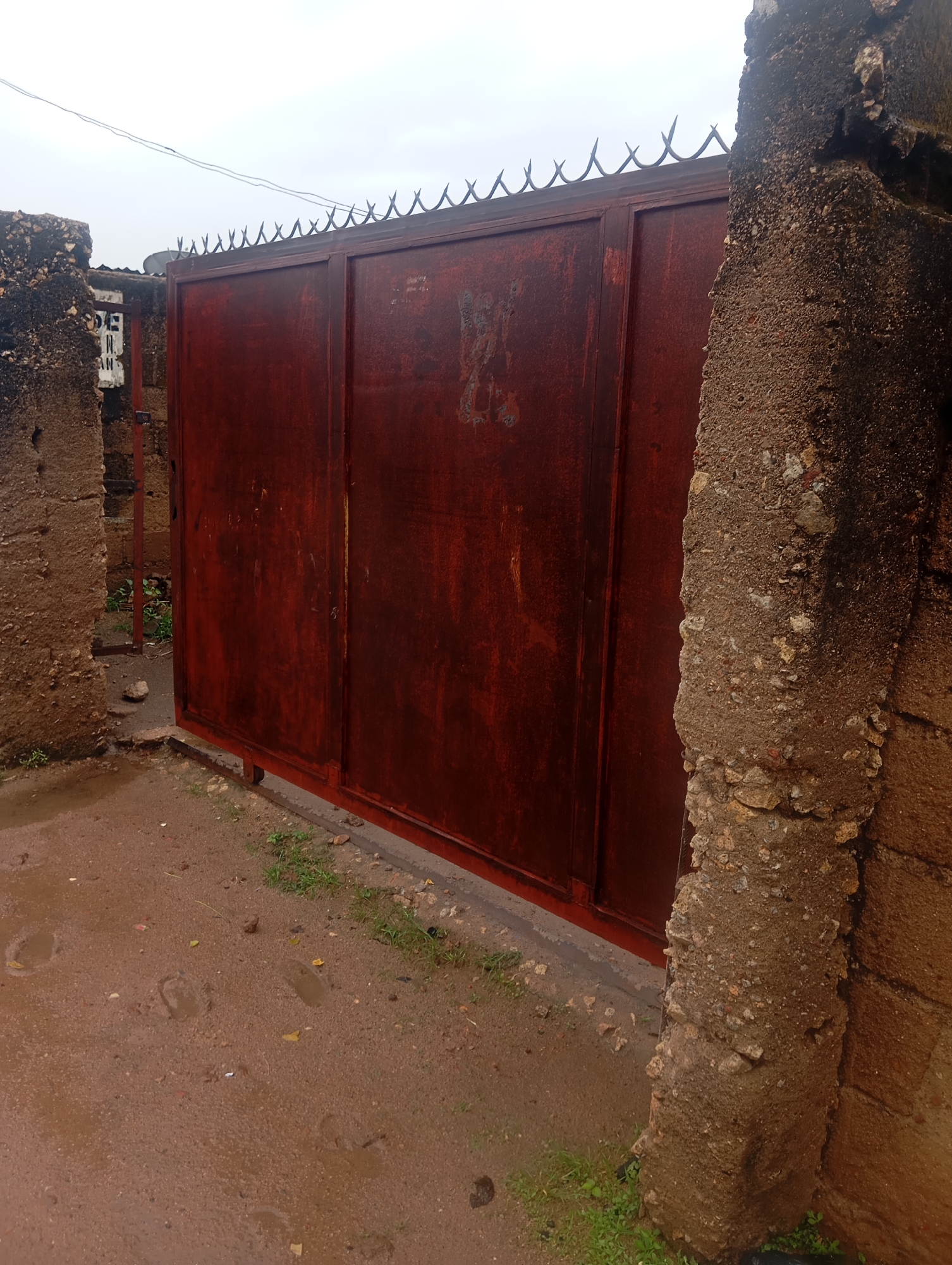 A wide, rusty red metal gate, approximately 10 feet tall, stands open, revealing a glimpse of greenery beyond. The gate is topped with pointed metal spikes, suggesting a security feature. It is flanked by two rough, concrete pillars; the one on the right is partially obscured by the gate itself and appears to be integrated into a wall. The ground in front of the gate is a muddy, reddish-brown dirt road, with several visible puddles and indentations that could be footprints. Some sparse green grass grows along the edges of the road and near the gate. The sky above is overcast and a pale gray. This scene is consistent with the entrance to a property in a residential or commercial area in Jos, Nigeria, where such gates and weathered construction are common.