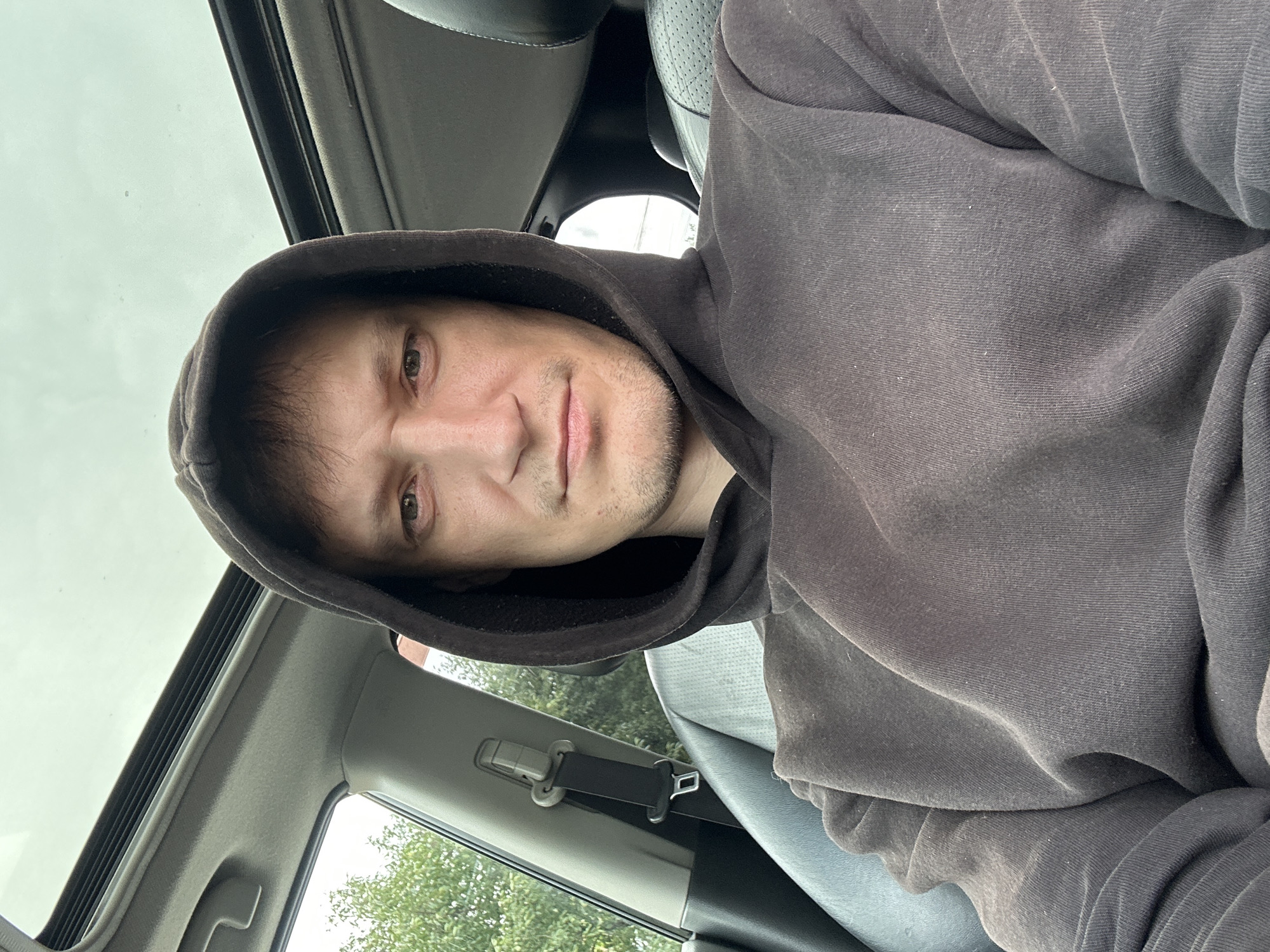 A man is taking a selfie in a car. He is wearing a dark grey hooded sweatshirt. The car has a sunroof, and the passenger seat and seatbelt are visible. The background shows trees and the sky. The image is a medium shot, focusing on the man's face and upper body.