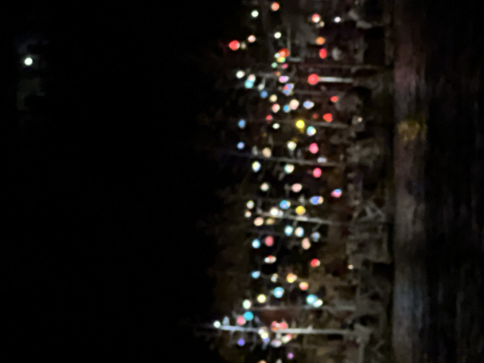 The image depicts a night scene with numerous colorful lights strung across trees or poles, creating a festive atmosphere. The lights are blurry due to the low light conditions and the camera's focus. In the foreground, the water reflects some of the lights, appearing dark and rippled. In the midground, there are indistinct shapes that suggest trees, structures, and possibly people seated in chairs or on benches. The background is entirely black, indicating a dark night sky. The overall impression is one of an outdoor nighttime event or celebration, possibly in a park or by a body of water.