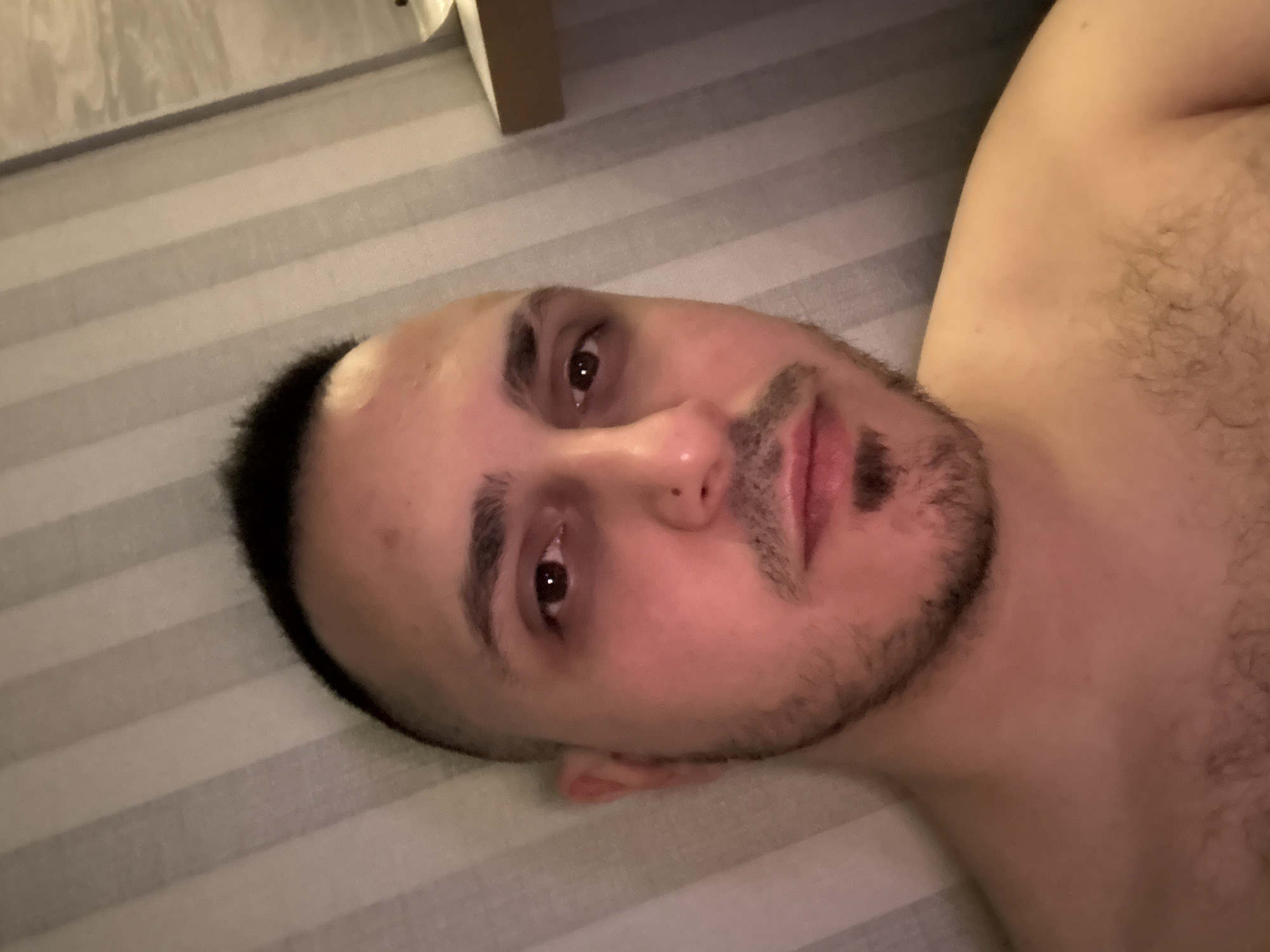 The media file contains a portrait of a man, oriented sideways, with his head positioned on the left side of the frame and his upper torso extending to the right. He has short dark hair, prominent dark eyebrows, brown eyes, and a short beard and mustache. His expression is neutral, and he is looking directly at the camera. There is a slight visible redness or darkness around his eyes. He appears to be shirtless, revealing some hair on his chest and shoulder. The background is a striped surface, likely a bedsheet or mattress, featuring alternating light and dark gray or beige horizontal stripes. In the upper background, there is a blurry glimpse of what appears to be furniture or a wall. The lighting suggests an indoor setting, possibly with artificial or dim light. The image captures a casual, close-up moment. There is no visible text.