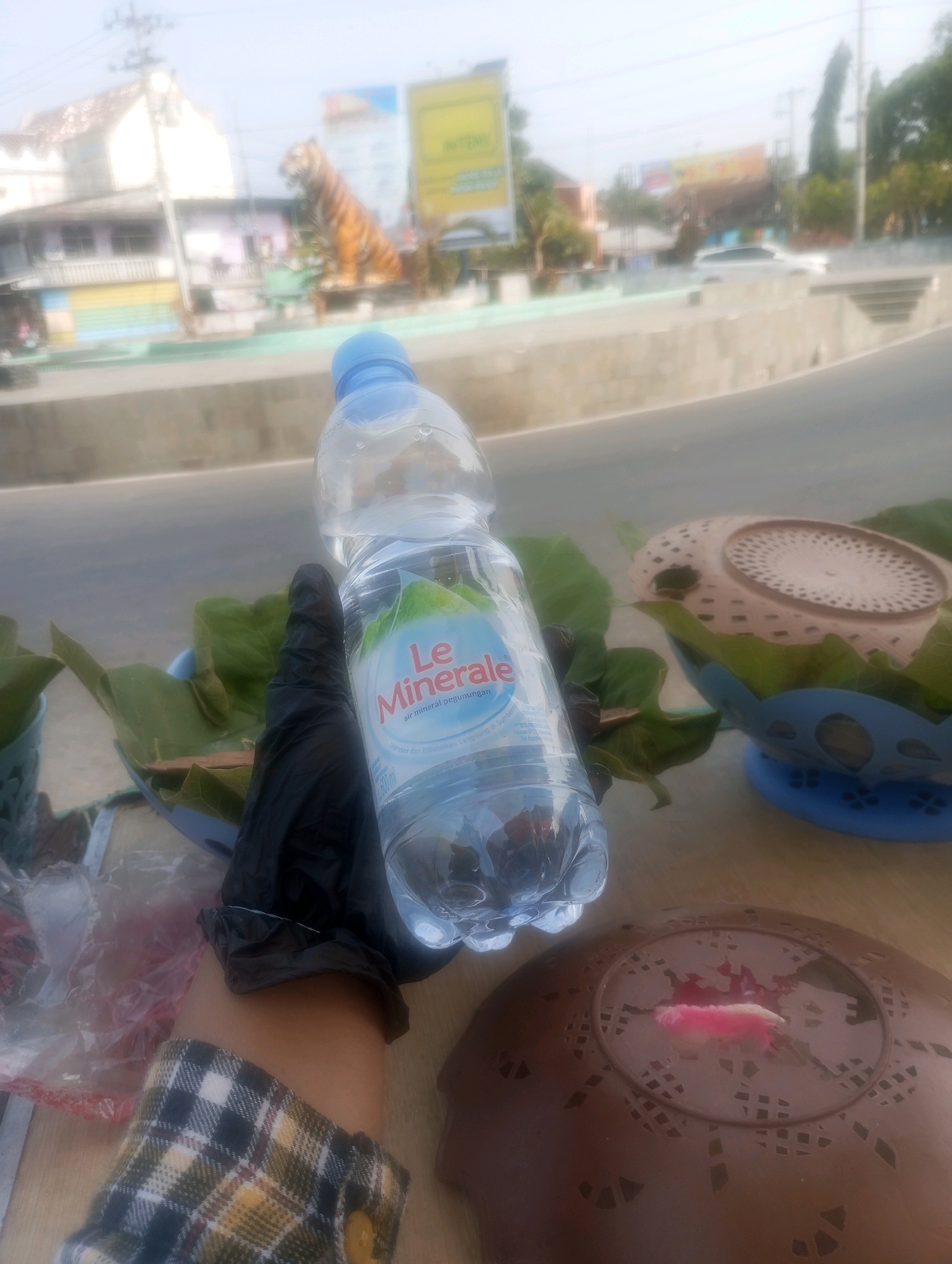 The image depicts a close-up view of a bottle of "Le Minerale" bottled water held by a person wearing a black glove, situated on a wooden surface. Behind the bottle, there are green leaves and a blue bowl containing what appears to be food wrapped in banana leaves. In the background, out of focus, there is a street scene with buildings, a tiger statue, and a car. The overall scene suggests an outdoor setting, possibly in a marketplace or roadside stall in Simo, Indonesia. The sunlight indicates it is daytime.