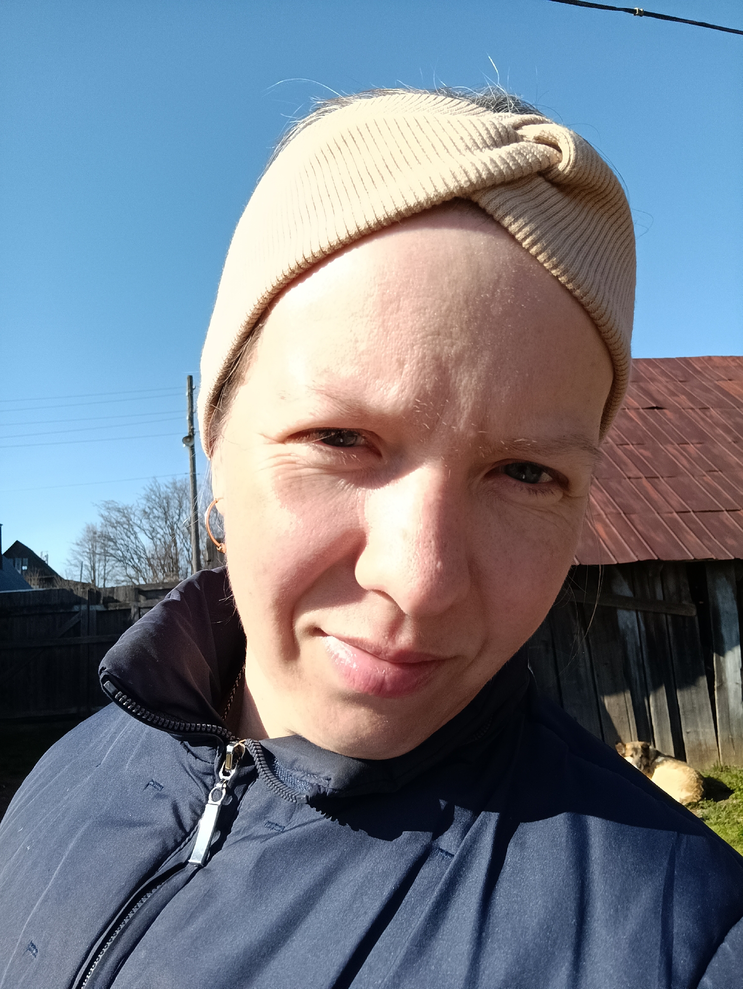 A medium-close-up shot shows a woman with fair skin wearing a beige knotted headband and a dark blue jacket. A silver zipper pull is visible on the jacket. She is smiling slightly. In the background, a wooden building with a rusty metal roof is present. A dog is lying in the grass next to the building. Trees and a blue sky are also visible in the background. The location is Пешнигорт, Russia.