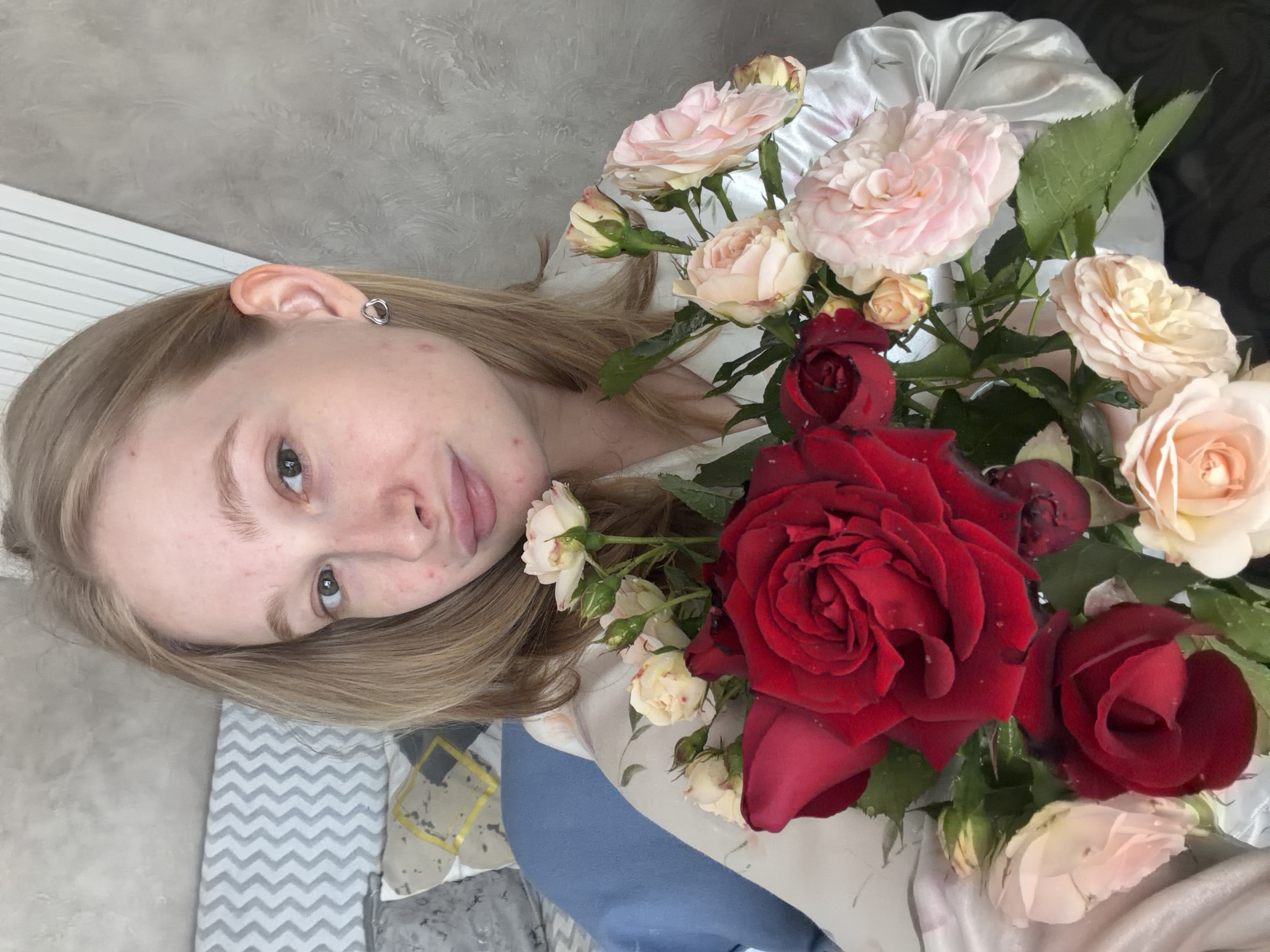 The image depicts a young adult female located in Rossiyskiy, Russia. She has shoulder-length blonde hair, light brown eyes, and fair skin with visible facial blemishes. She wears a small, silver-toned, irregularly shaped stud earring in her right ear. Her lips are lightly pursed, forming a subtle expression. She is dressed in a light-colored, possibly satin, garment with a faint floral pattern. She is holding a fresh bouquet of roses, featuring large deep red roses prominently, surrounded by smaller light pink and cream-colored roses and buds. Water droplets are visible on the petals and leaves. The background on the left includes a blue pillow, a grey and yellow patterned pillow, and a grey and white zigzag patterned fabric or headboard. The wall behind her on the right is light grey with a stucco texture. A white, vertically slatted structure, possibly a radiator cover, is visible in the upper right quadrant.