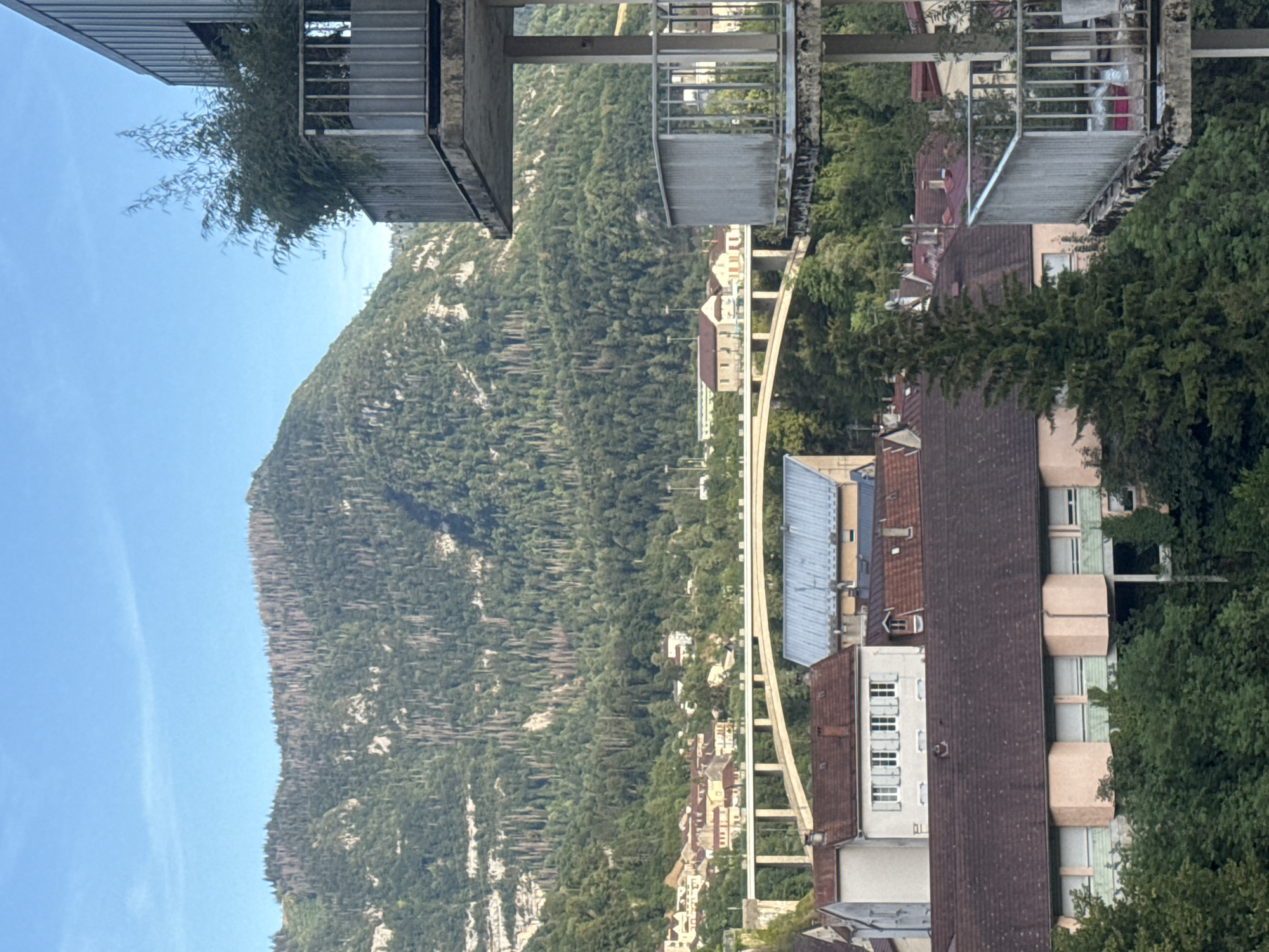 This image captures a view of the city of Saint-Claude, France, from a slightly elevated perspective, likely a building or balcony. In the foreground, a concrete balcony with a metal railing is visible on the right side, with some overgrown vegetation and a metal structure.
The midground features a cluster of residential buildings with terracotta roofs, typical of the region. Some buildings are lighter colored, with visible windows and architectural details like dormers. A prominent feature in the mid-background is a long, arched bridge spanning across a valley, connecting parts of the city.
The background is dominated by a large, densely forested mountain. The trees on the mountain are predominantly green, but there are also areas of sparser vegetation and some visible dead trees, particularly towards the summit. The mountain slopes are steep and rocky in places, with visible geological strata.
The sky is a clear, bright blue, indicating fair weather. The lighting suggests it is either late afternoon or early morning, given the soft light and the shadows cast. There are no people visible in the scene. The overall impression is one of a town nestled within a natural, mountainous landscape.