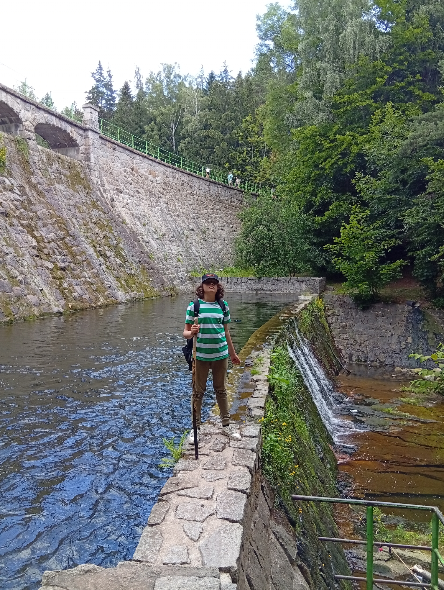 A person stands on a stone pathway between a body of water and a small waterfall. The person is wearing a green and white horizontally striped t-shirt, tan pants, white sneakers, and a dark baseball cap. They are holding a dark walking stick. Behind the person, a large stone dam is visible, with arched openings and a railing. Trees and foliage surround the dam and waterfall. The location is the Karpacz dam in Karpacz, Poland.