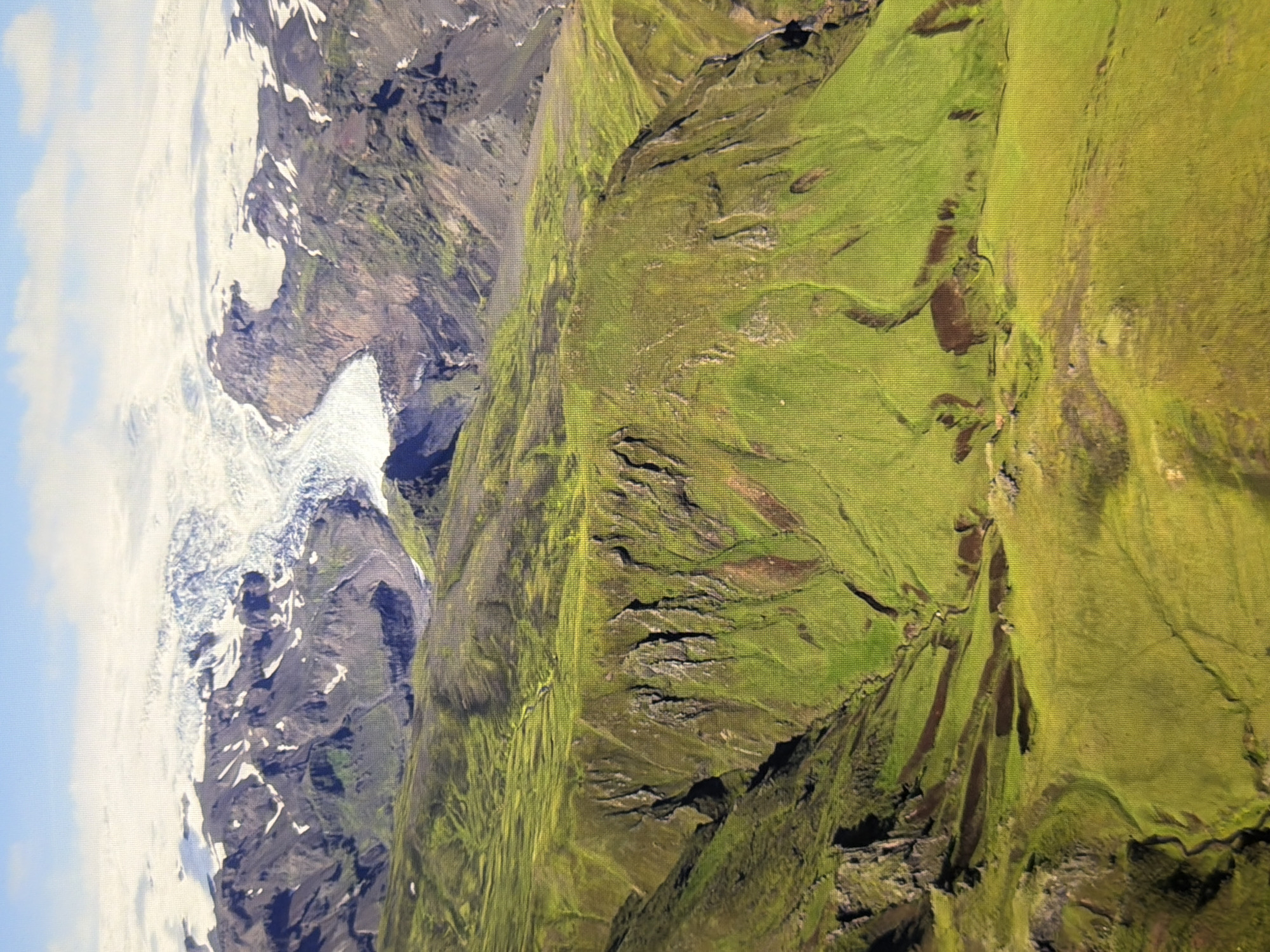 This is an aerial landscape photograph of a glacier in a mountainous region. The glacier is white and covers the top of the mountains. The mountains themselves are rocky and have patches of green vegetation. In the foreground, there are rolling green hills with dark patches that appear to be bodies of water or shadows. The sky is blue with some white clouds. There are no people or man-made structures visible in the image. The photograph is not related to the city of Kyiv, Ukraine.