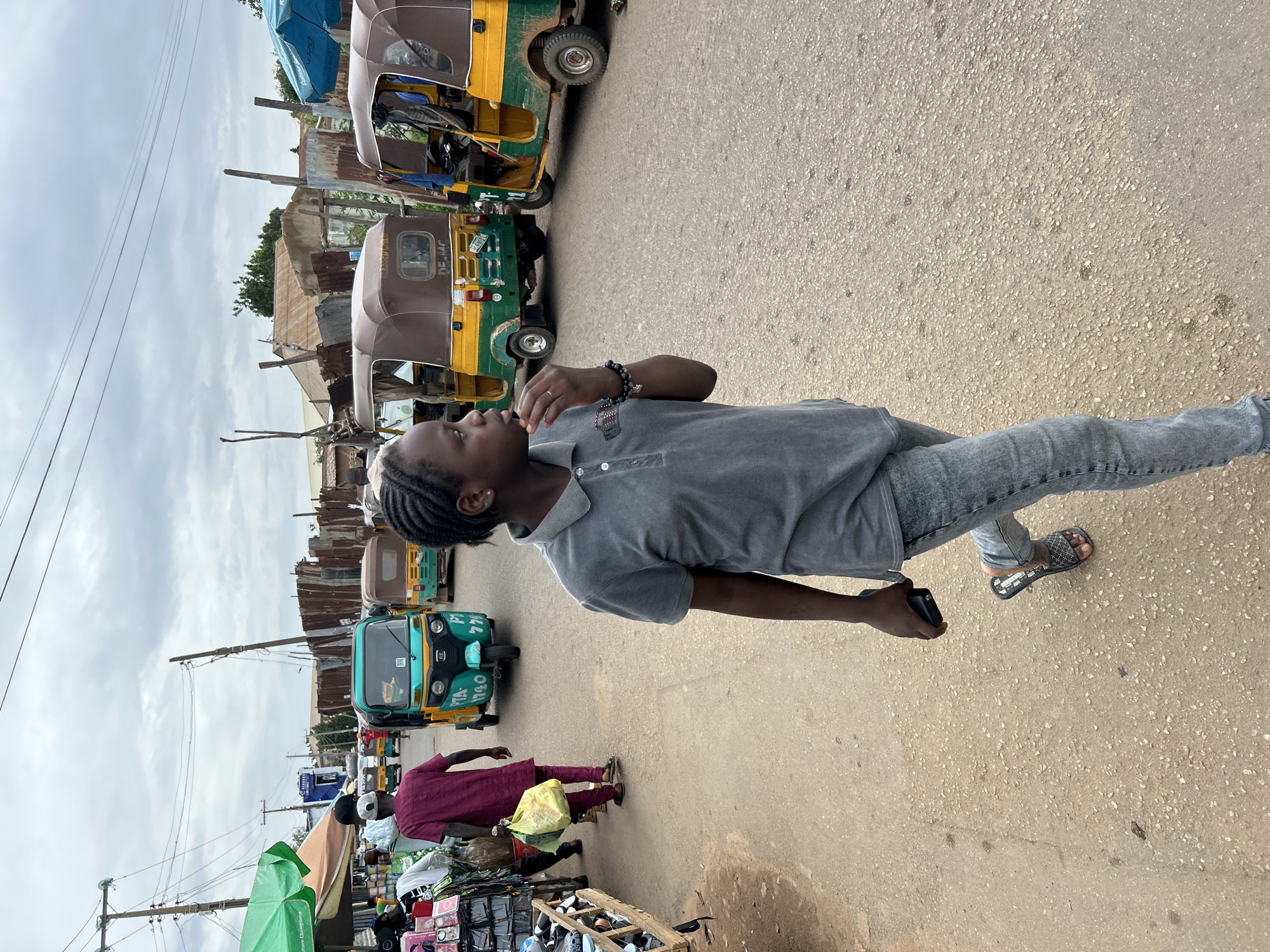 A person wearing a grey polo shirt and grey jeans stands on a paved street, holding a dark object in their right hand. They are facing to the left, with their head turned slightly. The person has braided hair and is wearing a bracelet on their left wrist. They are also wearing a sandal on their right foot.
The scene appears to be a busy street with several auto-rickshaws, also known as tuk-tuks, parked along the side. These vehicles are brightly colored, with green and yellow being prominent. In the background, there are buildings and structures that appear to be makeshift stalls or shops, some with corrugated metal roofs.
The weather appears to be overcast, with a cloudy sky visible. The lighting suggests it could be daytime, likely mid-morning or afternoon. The overall atmosphere suggests a market or a bustling urban environment, possibly in a developing country given the presence of the auto-rickshaws and the style of the buildings.
There is some visible text on the auto-rickshaws, including license plates and what appear to be registration numbers. For example, one vehicle has "DE-MJAC" visible. Another has "PTA-1190".
There are other people visible in the background, some walking and others engaged in activities related to the market stalls. One person in a maroon garment is carrying a yellow bag. The image is oriented vertically, with the subjects appearing from the bottom to the top of the frame.