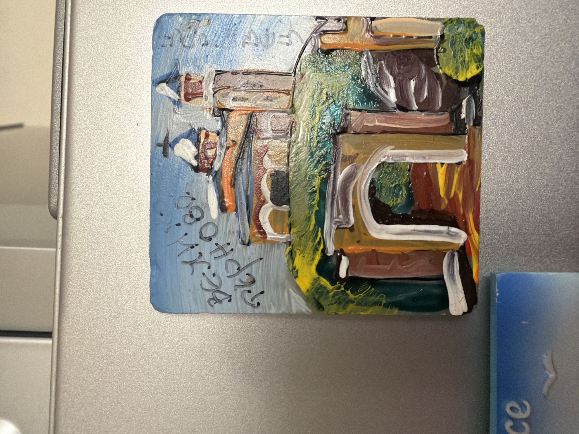 The image shows a magnet with a painting of a church on a hill. The church has a red roof and white towers. There is a blue sky in the background and some green trees. The painting is in a naive style, with thick brushstrokes. The magnet is attached to a metal surface, likely a refrigerator. The text on the magnet appears to be in Cyrillic and reads "В. Търново", which translates to "Veliko Tarnovo". Veliko Tarnovo is a city in northern Bulgaria, known for its historical significance and picturesque location.