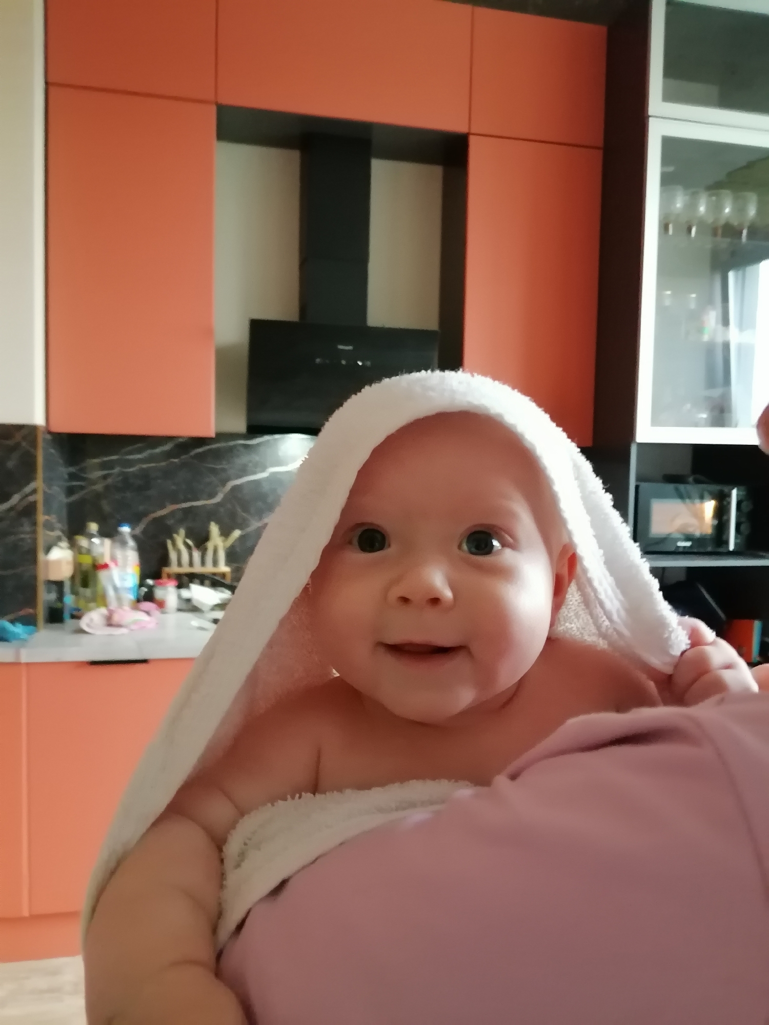 A baby, wrapped in a white towel, is being held by an unseen person. The baby is looking at the camera with a slight smile. The background appears to be a kitchen with orange cabinets. The location is in the city of Trubacheyevka, Russia.
