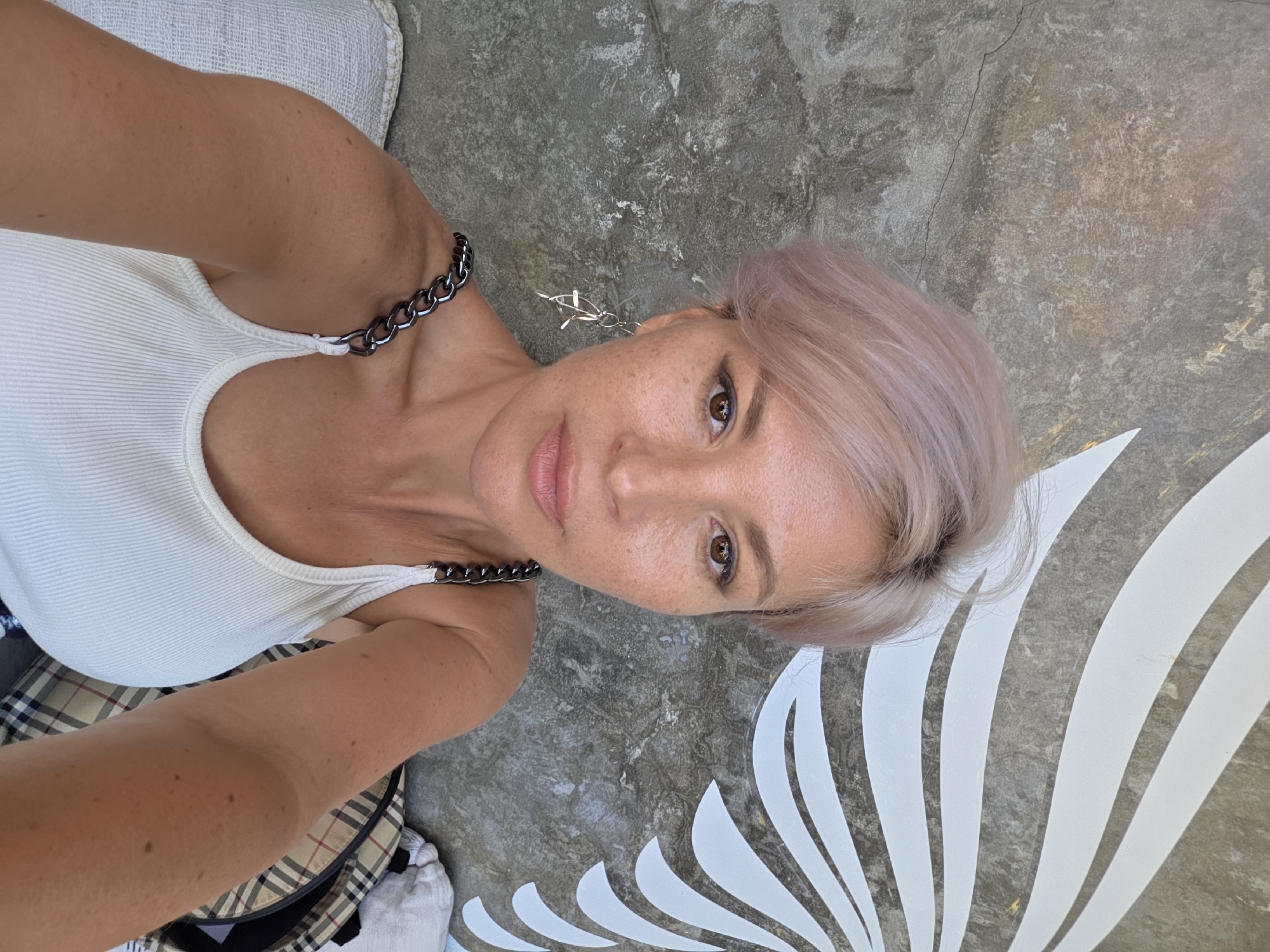 A woman is taking a selfie outdoors. She is wearing a white ribbed tank top with chain straps and plaid pants. Her hair is short and light pink. She is standing in front of a concrete floor with a white decorative design painted on it. The location is likely North Kuta, Indonesia, based on the environment and style of the image.