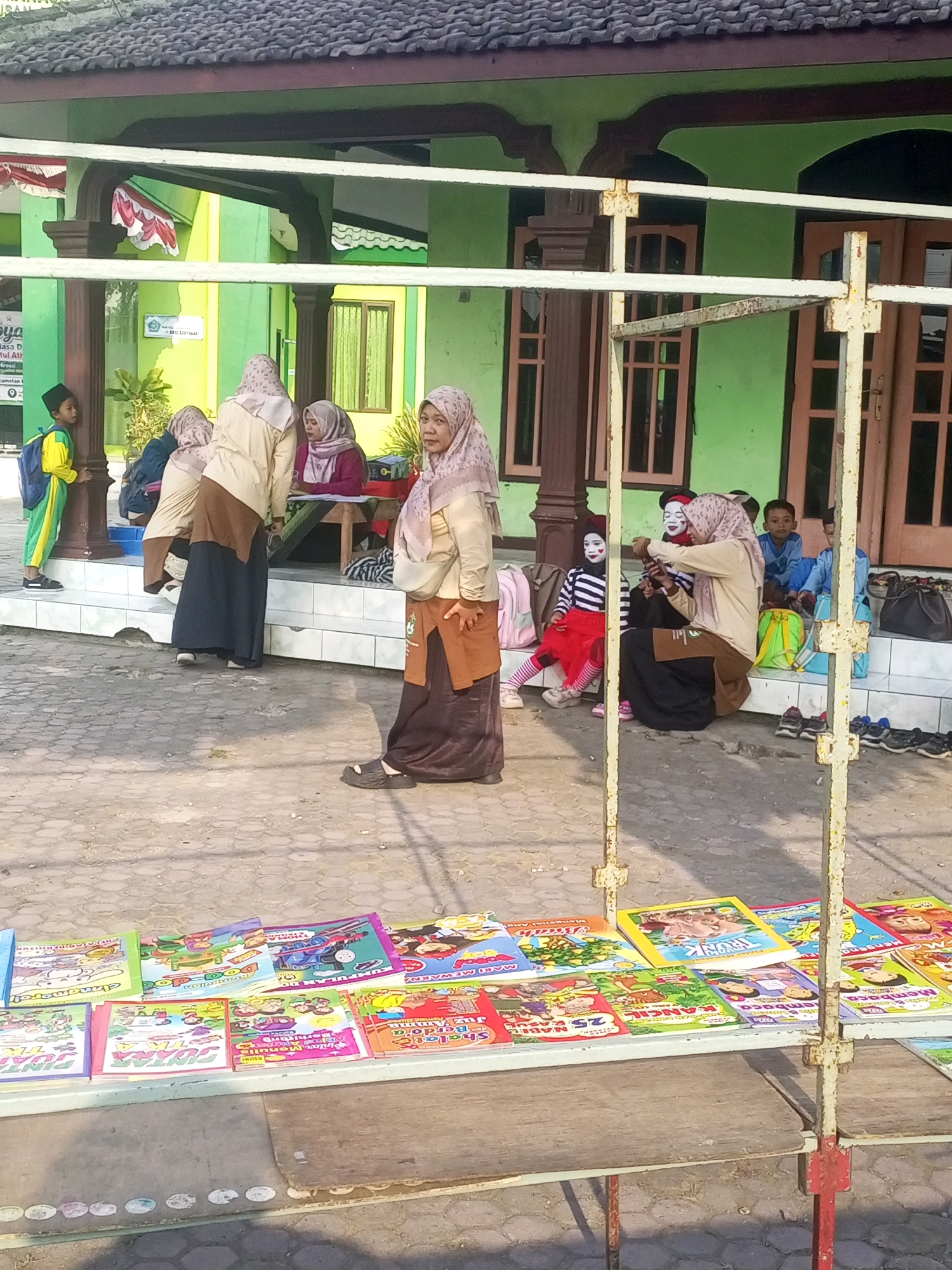 The image depicts an outdoor scene at what appears to be a school or community event in Gurah, Indonesia. In the foreground, a table displays a variety of colorful children's books. Behind the table, several individuals are present. A woman wearing a hijab and brown skirt stands in the center, looking towards the left. To her right, another woman in a hijab is seated at a table, seemingly interacting with a child who has face paint resembling a clown. Other children and adults are visible in the background, some seated on steps and others standing. The environment includes a building with green walls and a dark tiled roof, along with a metal structure that supports the table of books. The overall atmosphere suggests a casual gathering, possibly related to educational activities or a local market.