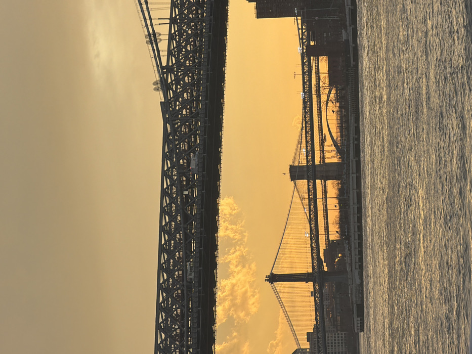 The image displays a waterfront scene on the East River in New York City, United States of America, during golden hour, characterized by a predominantly golden-orange sky. The upper section is dominated by the Williamsburg Bridge, a steel truss bridge with multiple light-colored vehicles discernibly moving on its upper deck. Below this, the Manhattan Bridge, a suspension bridge with two prominent towers and visible deck lights, is positioned to the left. To the right of the Manhattan Bridge, the Brooklyn Bridge, another suspension bridge featuring a distinct tower and illuminated vehicle traffic, is visible. Additional lower bridge segments and an arched structure are present beneath and behind these main bridges. The lower third of the image depicts the rippled surface of the East River, reflecting the sky's warm tones. The distant landmasses feature silhouetted urban buildings, with a large, dark building structure visible on the far right shore. No individual human figures are distinctly identifiable.