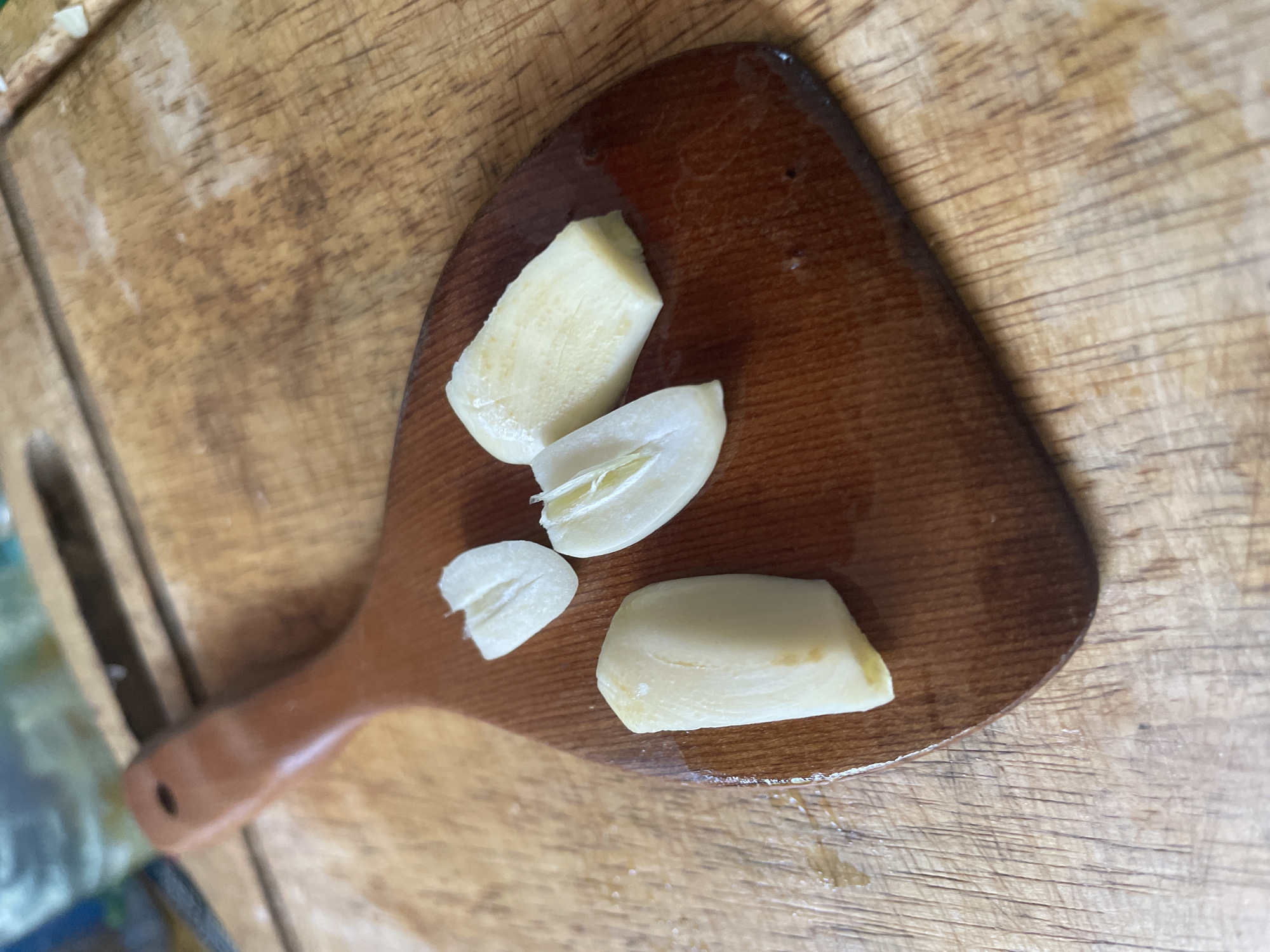 A wooden spatula with four pieces of peeled garlic on it sits on a wooden cutting board. The cutting board appears to be well-used, with visible knife marks and a slightly worn surface. The spatula has a dark brown, lacquered finish and a hole at the end of its handle. The garlic pieces vary in size and shape, with some being whole cloves and others cut into smaller segments. The scene is lit by natural light, casting soft shadows. The background is slightly blurred, suggesting a kitchen or food preparation area. This image does not provide any specific geographical information about Phu Thuy Ward, Vietnam.