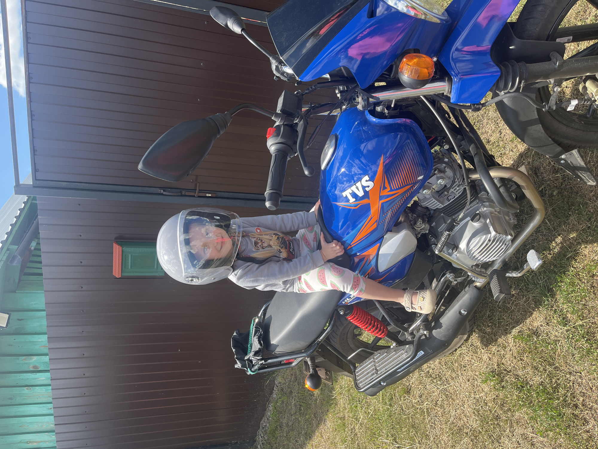 A young girl wearing a clear-visored helmet sits on a blue motorcycle parked on a grassy area in front of a brown metal fence. The girl is wearing a grey long-sleeved shirt with a cartoon image and patterned leggings. The motorcycle has orange accents and the "TVS" logo on the fuel tank. In the background, a green wooden structure with a small red and green mailbox is visible. The scene appears to be in a rural or suburban setting, possibly in the city of Сухринское, Russia, given the context. The sky is bright blue with some clouds. The ground is covered in dry grass and some patches of green vegetation.