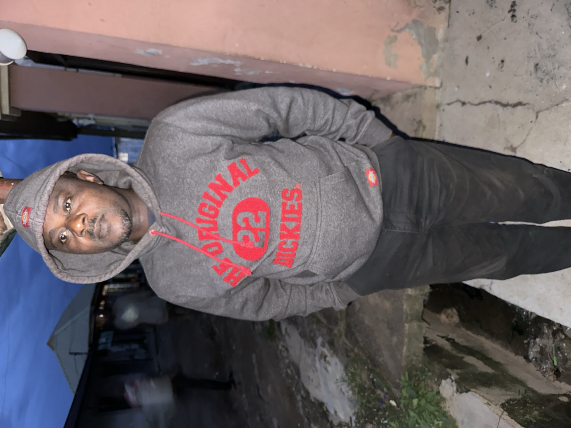 In the city of Narayi, Nigeria, a man stands outdoors at dusk. He is wearing a grey hooded sweatshirt with red lettering that reads "THE ORIGINAL 22 DICKIES". The man has dark skin and is looking directly at the camera. He is standing in what appears to be a residential or commercial area, with buildings visible in the background. There is a drain or ditch in the foreground. The sky is dark blue, indicating the late hour.