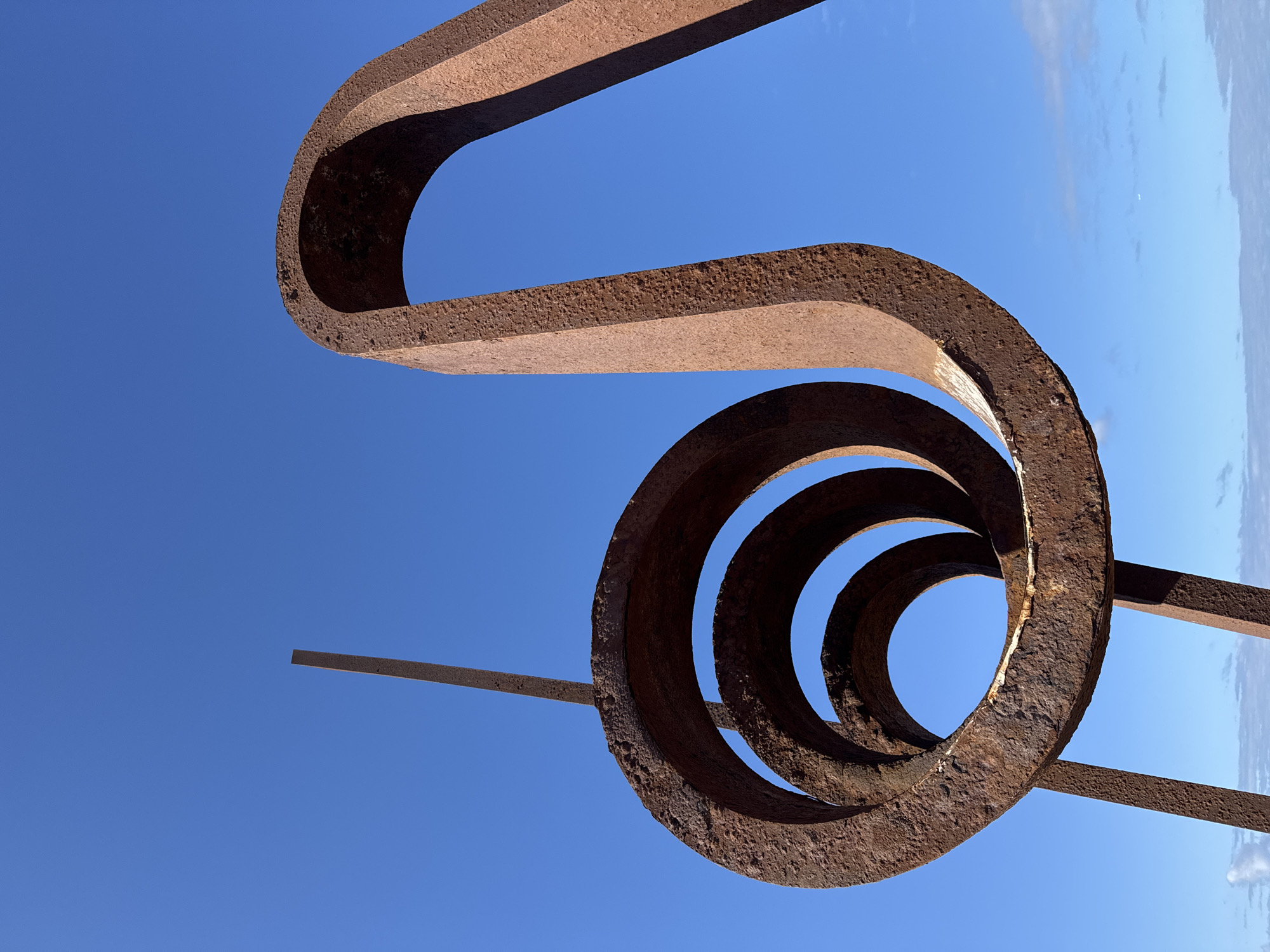 A low-angle outdoor shot captures a large, rusted metal sculpture against a clear blue sky. The sculpture consists of two distinct parts: on the left, a series of concentric circles, each with a progressively smaller diameter, are stacked and appear to be formed from a coiled metal band. A thin, vertical metal rod extends upwards from behind these circles. On the right, a thick, U-shaped metal beam curves upwards, forming an arch. The metal of the sculpture is weathered, showing signs of rust and texture. The background is dominated by a bright, cloudless blue sky, with a sliver of the ocean and a distant horizon visible at the bottom of the frame. There are no people or animals visible in the image. The sculpture is located on the Playa de Las Américas, Spain.