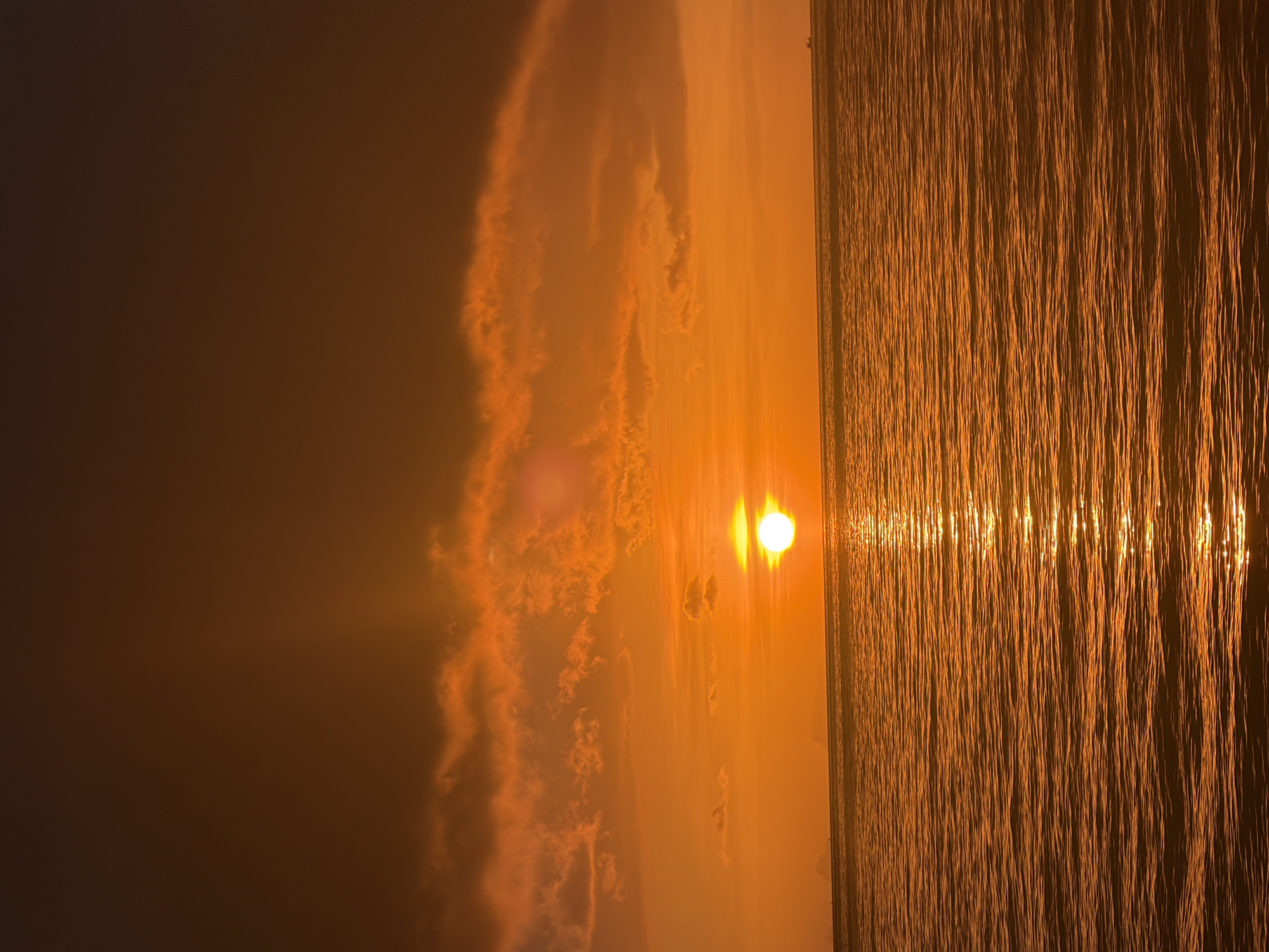 The image depicts a sunset over the sea in Baan Tai, Thailand. The sun, a bright orb, is positioned in the center-right of the frame, casting a golden glow across the water and illuminating scattered clouds. The water is a deep orange, with ripples reflecting the sunlight. In the distance, faint silhouettes of landmasses or islands are visible on the horizon. The sky transitions from a bright orange near the horizon to a dark, reddish-brown at the top of the frame. A small boat is visible on the horizon in the far right.