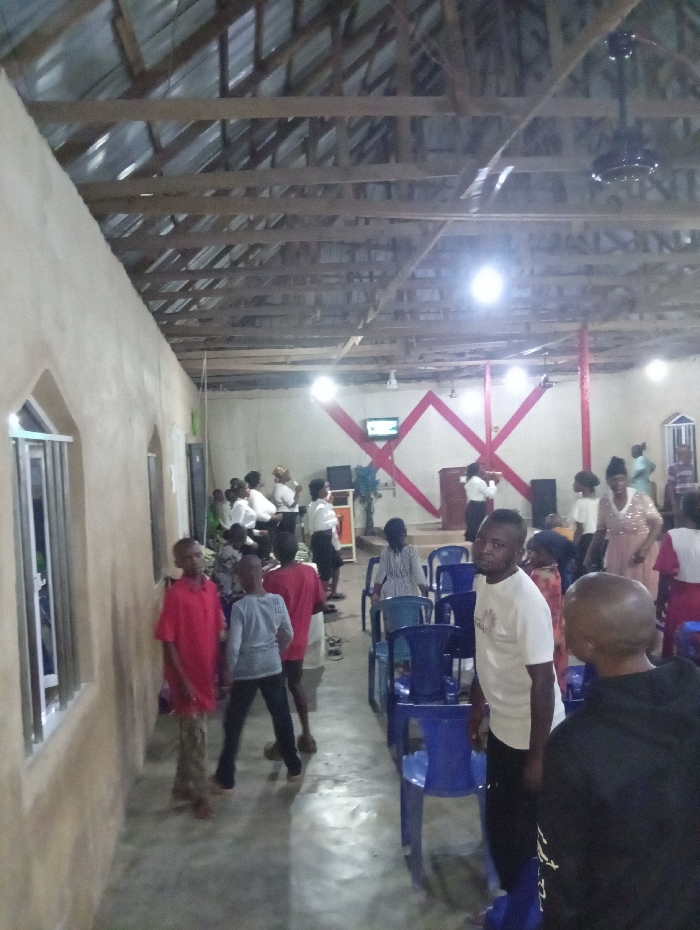 An indoor shot shows people gathering in what appears to be a church or meeting hall. The hall is minimally furnished with blue plastic chairs, and the walls are plain. Some individuals are standing or walking near the entrance area on the left, while others are congregated closer to a raised area at the back, where a television and red decorative elements are visible. The ceiling is exposed with wooden beams and a corrugated metal roof. The lighting is provided by bright overhead fixtures.