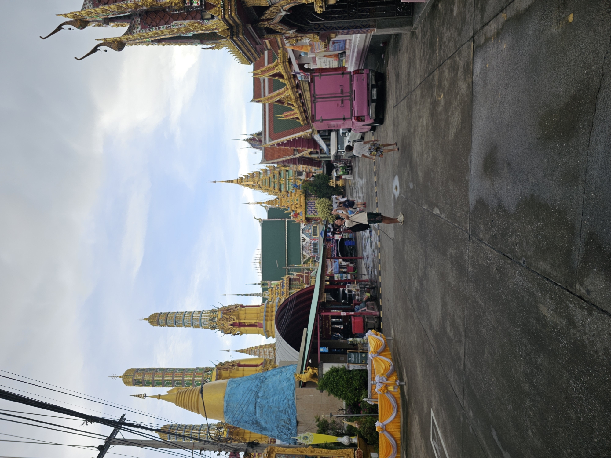 The image depicts a view down a street lined with ornate Thai temples and structures, likely in Bangkok, Thailand, given the architectural style. The foreground shows a paved street with some puddles, indicating recent rain. On the left, a vibrant yellow and white draped fabric is set up, possibly for an event. Further down the street, several towering, multi-tiered structures with gold and colorful tiled spires are visible, characteristic of Thai temples. A large, blue, conical structure is also prominent on the left.
In the middle ground, a pink truck is parked on the right side of the street. Several people are present; one man in the middle ground on the left is taking a photo with his phone, wearing a white t-shirt and shorts. Another person, a woman, is walking away from the camera on the right, wearing a patterned dress. Other individuals are seen in the distance, some near food stalls or shops that line the street. The sky is overcast with clouds. The overall scene suggests a bustling area with religious and commercial activity.