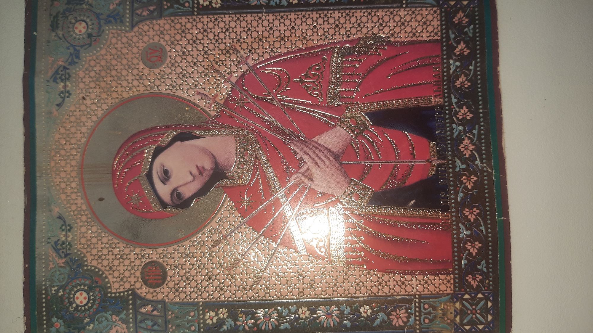 The image is a close-up, slightly angled, horizontal shot of an Eastern Orthodox icon. The icon depicts the Virgin Mary, shown from the chest up, with her head tilted and eyes looking slightly down and to the left. She is wearing a red robe adorned with intricate gold detailing and appears to be pierced by seven swords or arrows, arranged around her chest. Her hands are clasped over her chest. The background of the icon is a patterned gold foil with a circular halo around Mary's head, also outlined in gold. The icon is framed by ornate borders featuring floral and geometric patterns in dark colors, gold, and light blue. The overall condition of the icon appears to be somewhat worn, with visible creases and some discoloration, suggesting age. There is no discernible information within the image that would indicate its specific location in the city of Shklov, Belarus.