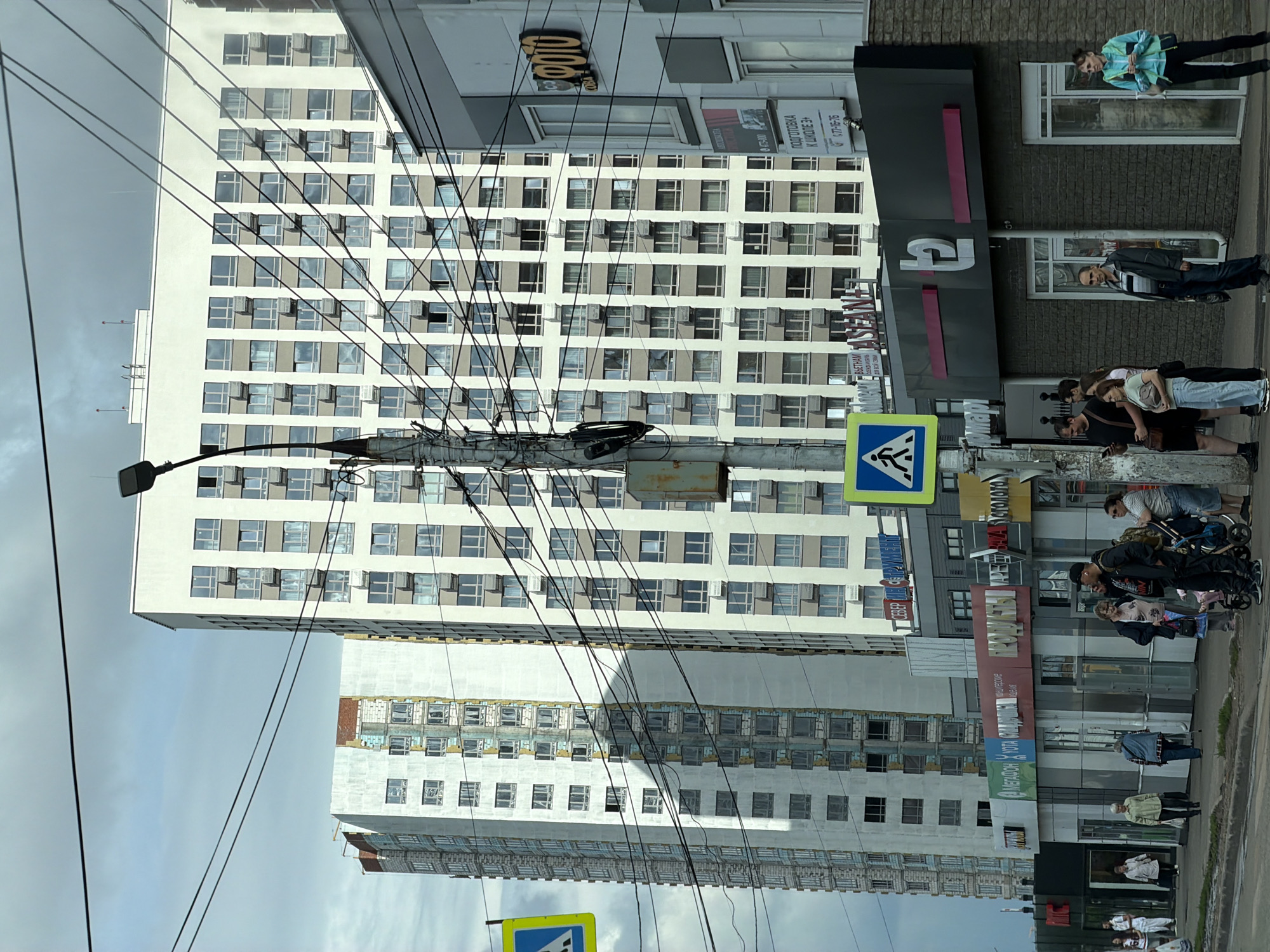 The image depicts a street scene in an urban environment with several multi-story buildings. The foreground features a sidewalk with a group of people standing and walking. A street light pole with wires is prominently positioned in the center. The buildings in the background are tall residential structures with numerous windows. Some buildings appear to be under construction or renovation, with exposed brickwork visible on one. A blue and yellow pedestrian crossing sign is visible on the left side of the image. Storefronts with signage are present at the ground level of some buildings. The sky is overcast, suggesting cloudy weather.