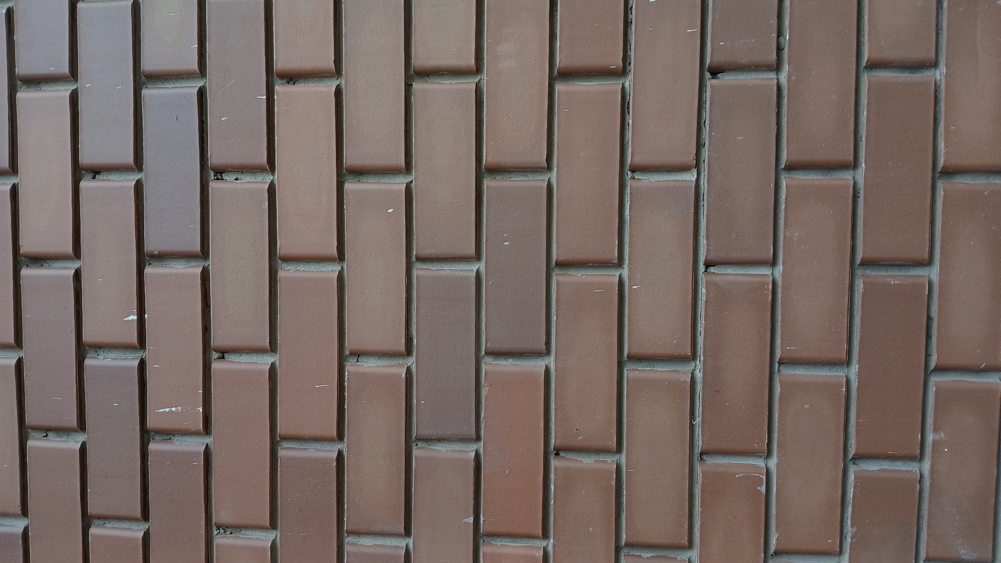 The image is a close-up, vertical shot of a brick wall. The bricks are rectangular and have a reddish-brown hue with some variations in color, suggesting a natural clay material. The mortar between the bricks is a lighter gray color. The bricks are laid in a staggered pattern, a common bricklaying technique. There are some faint white marks visible on a few of the bricks, possibly dust or scuff marks. The lighting is even, suggesting an overcast day or an indoor setting with diffuse lighting. The image provides no information about the city of Oral, Kazakhstan, or any specific location within it.