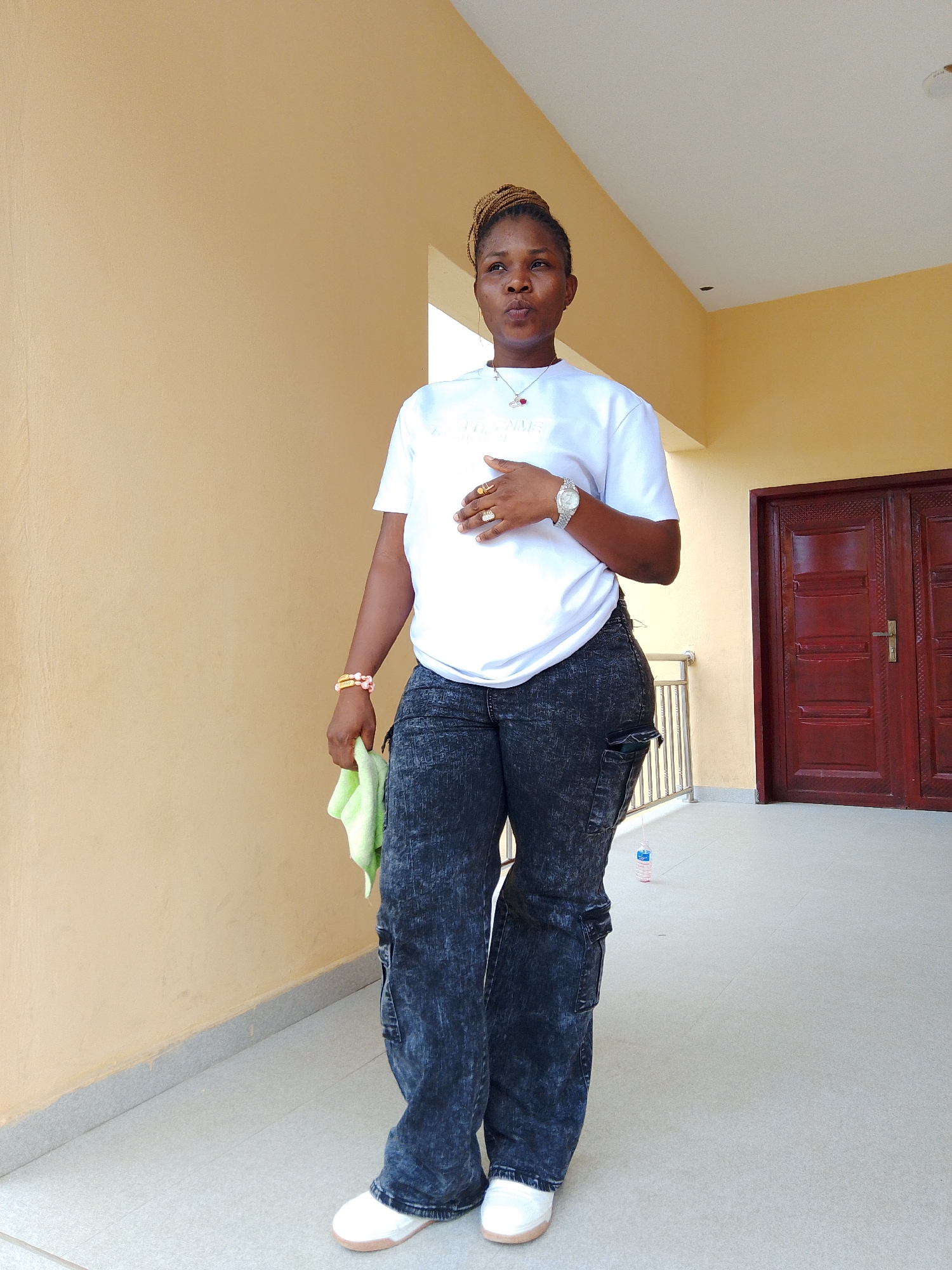 A full-body, slightly low-angle shot shows a Black woman standing on a light-colored tiled walkway. The woman is wearing a white t-shirt with a graphic design, dark-wash, acid-wash cargo pants, and white slip-on shoes. She has her left hand on her chest and her right hand holding a light green cloth. She is also wearing jewelry, including rings on her fingers, a watch on her left wrist, a necklace with a red pendant, and bracelets on her right wrist. Her hair is styled in braids that are pulled back.
Behind the woman, a wall with a textured, light yellow finish runs the length of the walkway. To the right, a large, dark red double door with intricate carvings is visible. A metal railing runs along the edge of the walkway. A plastic water bottle with a pink label is on the ground near the railing.
The scene appears to be an outdoor area, possibly a veranda or hallway of a building, with the warm tones of the wall and the contrasting dark door suggesting a possibly commercial or residential setting in Yenegoa, Nigeria. The lighting suggests it is daytime.
