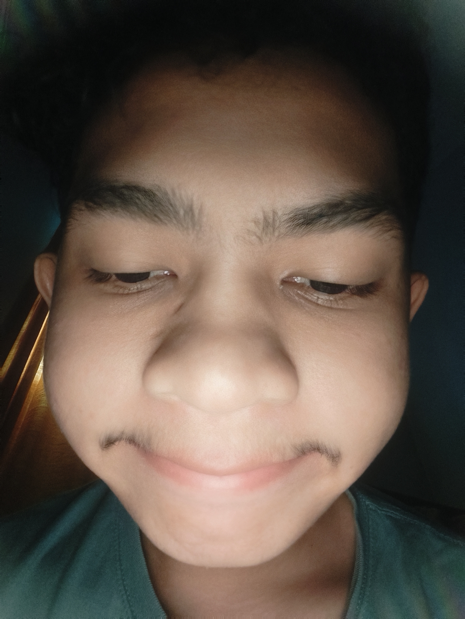 The image displays a close-up, frontal view of a single individual's face, occupying the majority of the frame. The subject, a young person with light brown skin, features dark eyebrows and dark hair partially visible at the top. Both eyes are oriented downwards. The cheeks are visibly puffed out. A faint, sparse dark line, consistent with a nascent mustache, is present above the upper lip. The mouth is closed with a subtle upward curve. The individual is wearing a teal-colored garment, visible across the lower portion of the image. The lighting is focused on the face, with the background remaining indistinct and dimly lit. The scene is located in Lhokseumawe, Indonesia.