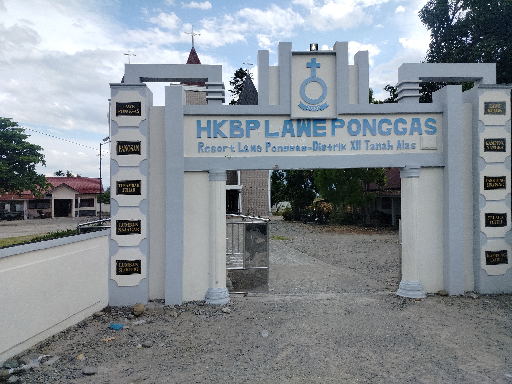 The image shows an entrance archway to a religious institution, identified by the text "HKBP LAWEPONGGAS Resort Lake Ponggas-Distrik XII Tanah Alas" above the arch. The archway is constructed from light-colored concrete with blue accents. A cross is visible on top of a red-roofed building behind the arch, and another cross is mounted on a dark-colored conical spire also behind the arch. There are several black signs with white lettering attached to the pillars of the archway, listing place names such as "LAWE PONGGAS", "PANOSAN", "TENAMBAK JUHAR", "LUMBAN NAJAGAR", "LUMBANT SITIO", "LAWE KULOK", "KAMPUNG NANGKA", "TARUTUNG SINAPANG", "TELAGA TUJUH", and "KAMPUNG BARU". The ground in front of the archway is covered in gravel and dirt, with some scattered debris including plastic bags and stones. To the left of the arch, there is a white wall and a building with a red roof. Behind the arch, a paved pathway leads into the compound, where a gate made of metal bars is visible. Trees and other vegetation are present in the background. The sky is partly cloudy with a bright, overcast daylight. The scene appears to be in a rural or semi-rural area, likely within Indonesia, given the signage.
