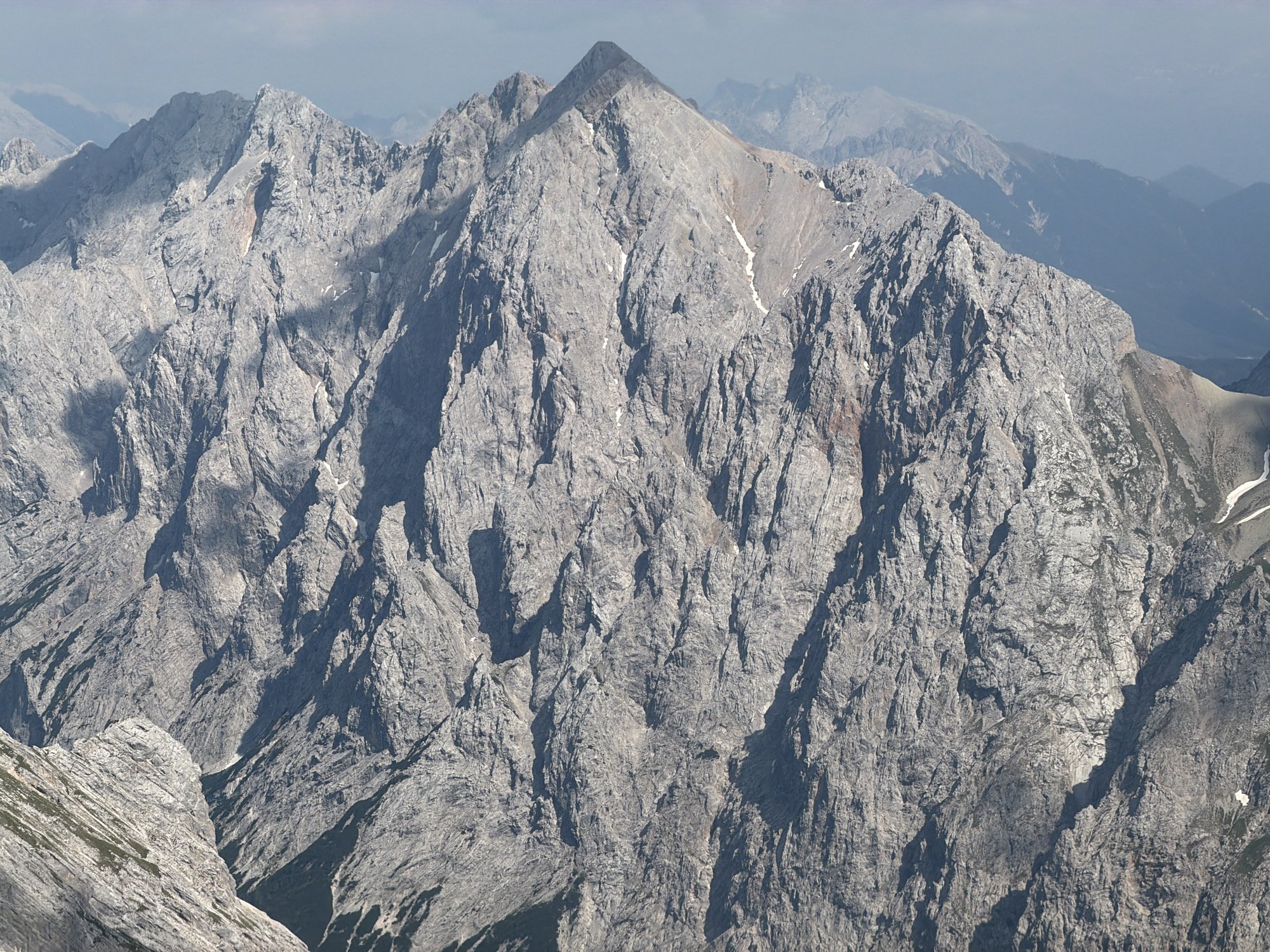 This media file captures a majestic, awe-inspiring mountain landscape under a partially cloudy sky, suggesting either early morning or late afternoon lighting. The scene is dominated by rugged, grey, rocky peaks with deep ravines and steep slopes. Patches of snow are visible on some of the higher elevations, hinting at a cooler climate or season. The textures of the rock faces are detailed, showcasing erosion and natural formations that have shaped these mountains over time. In the background, successive layers of mountains fade into a hazy blue, indicating a vast and expansive mountainous region. The overall mood is one of solitude and the raw power of nature. There are no people or man-made objects visible, emphasizing the wild and untouched character of the location. The lack of specific landmarks makes precise location pinpointing difficult without external information, but the described topography is consistent with alpine environments. The time of day is suggested by the shadows cast across the mountainsides, with some areas brightly illuminated and others in deep shadow, creating a dramatic contrast. The weather appears to be fair with clouds contributing to a dynamic sky rather than indicating immediate precipitation. The absence of any visible text further reinforces the natural, unadulterated subject matter of the image.