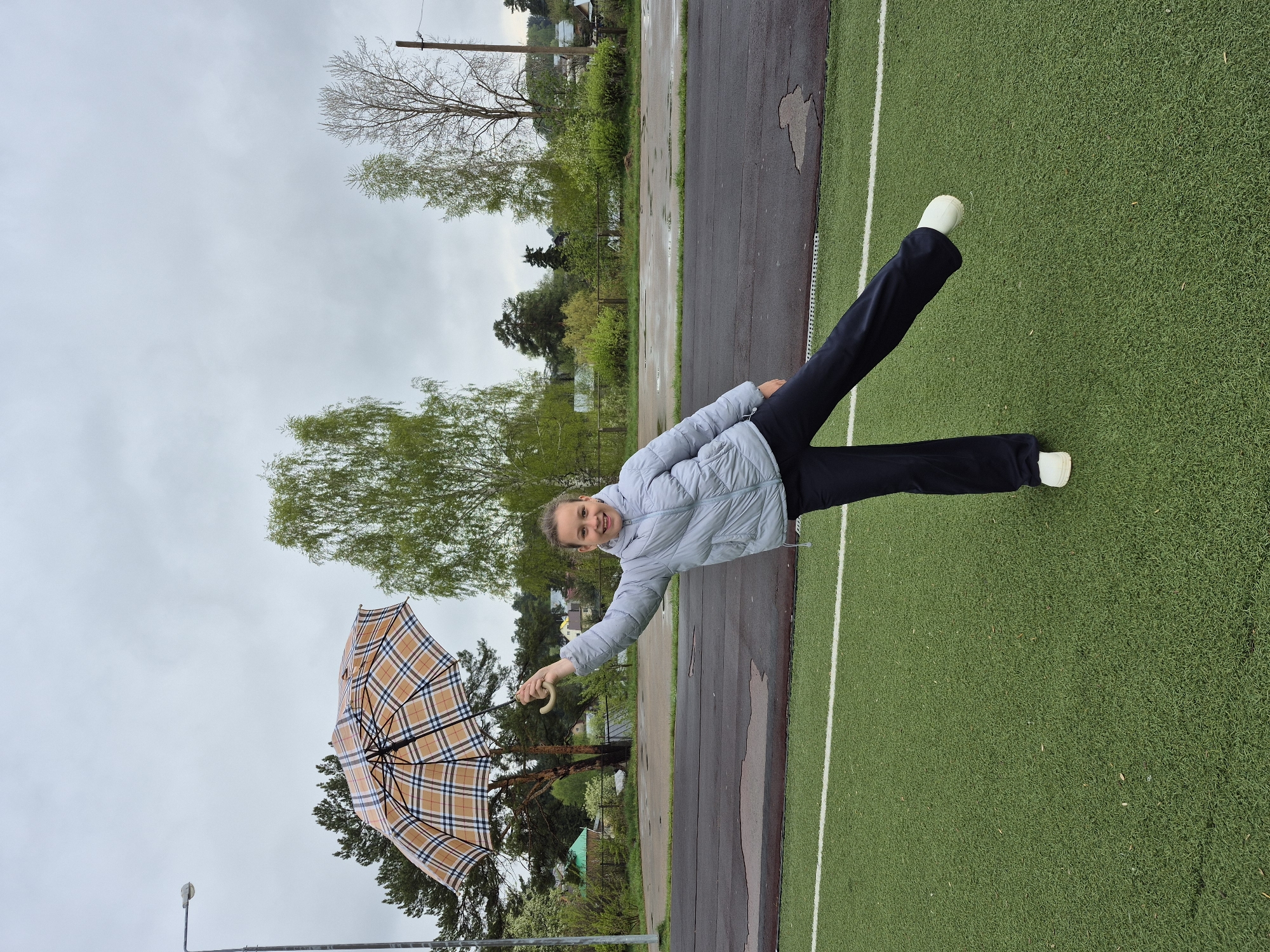 The image depicts a woman lying horizontally on a green surface that appears to be artificial turf. She is wearing dark pants, white shoes, and a light blue quilted jacket. She is holding a tan and plaid umbrella with a curved handle. The scene seems to be taken at a track field with a paved running track adjacent to the turf. In the background, there are trees and a building, suggesting an outdoor location in a park or recreation area. The city of Chaikovsky, Russia, is likely the location, although this cannot be definitively confirmed from the image alone. The sky is overcast. The woman is looking at the camera and smiling.
