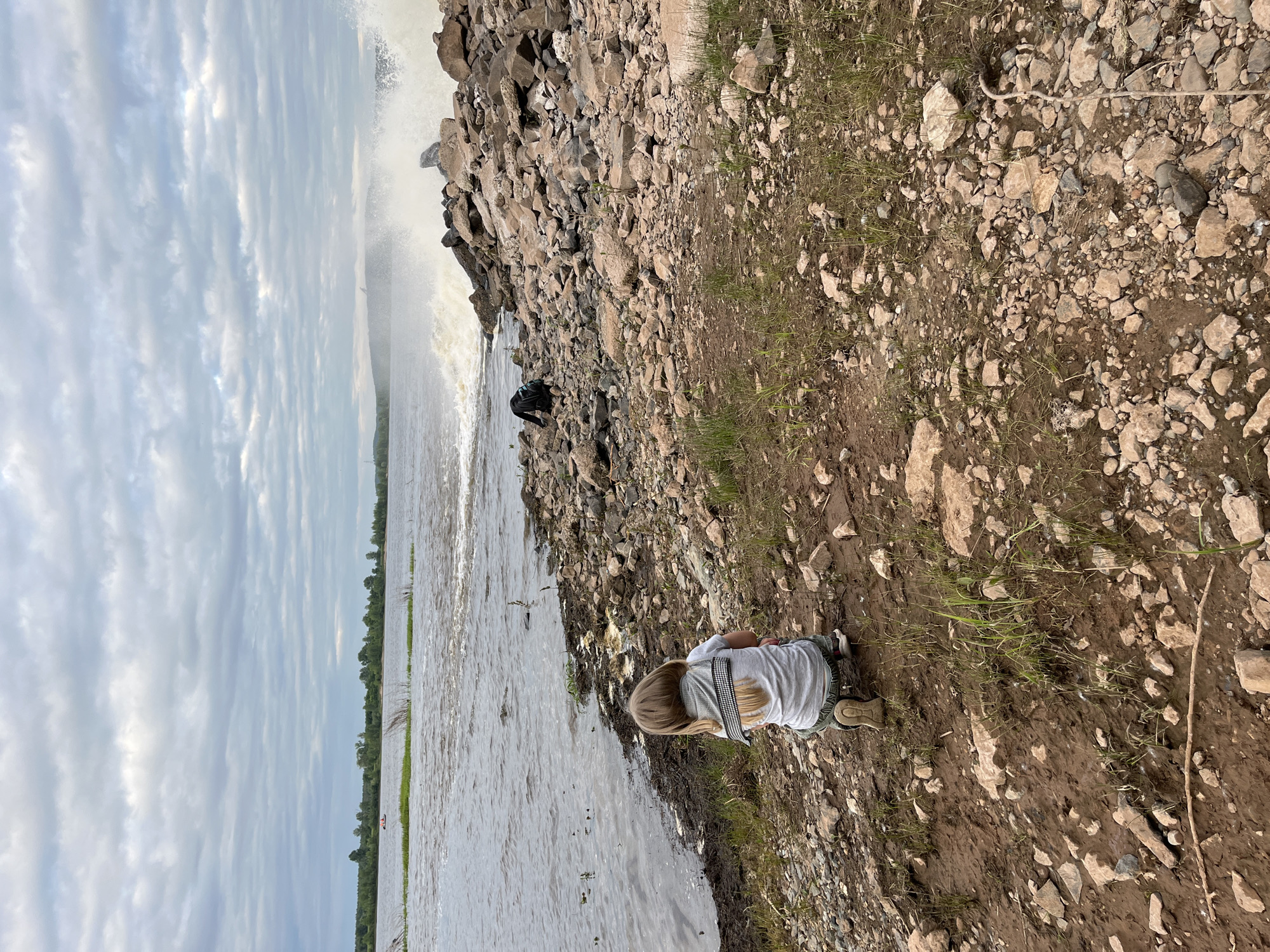 A young child with blonde hair, facing away from the camera, kneels on a rocky, muddy shoreline next to a large body of water. The child is wearing a light-colored t-shirt and dark pants. In the background, waves crash against a rocky embankment. The water appears choppy, and the sky is overcast with clouds. A backpack is visible near the embankment. The scene is located on the shore of the Ufa River in the city of Chernushka, Russia.