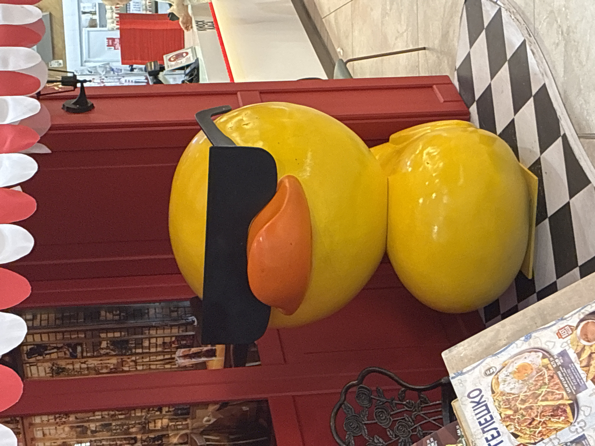 A large, yellow, stylized duck statue wearing black sunglasses and an orange beak stands on a black and white checkered floor. The duck is positioned in front of a red building facade with a red and white striped awning. To the left of the statue, a menu on a stand displays pictures of food, and a decorative black metal chair is visible. In the background, a white counter with a red accent light is present, and a person is partially visible on the right side. The overall setting appears to be an outdoor seating area or a storefront in a commercial district. This scene is located in Sofia, Bulgaria, specifically within the mall 'City Center Sofia'.