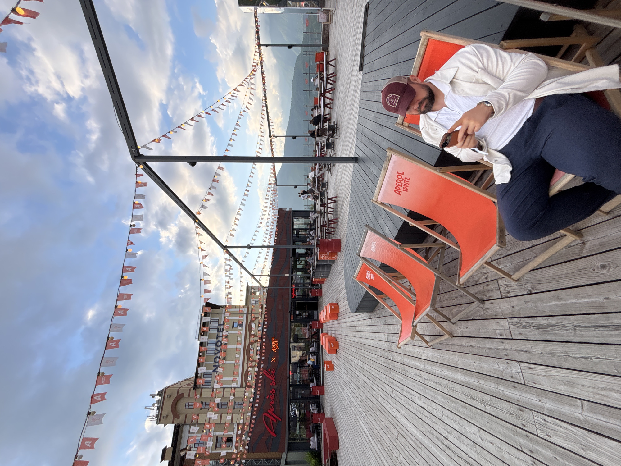 The image captures an outdoor scene on a wooden deck, likely during daylight hours given the bright sky. The sky is a mix of blue with fluffy white clouds, suggesting fair weather. In the foreground, a man is seated in a bright orange deck chair, holding a smartphone. He is wearing a white shirt, a light-colored jacket, and a maroon baseball cap. Another identical orange deck chair is visible next to him.
The background reveals a bustling atmosphere. Rows of tables and chairs are set up, with several people seated and interacting, indicating a social gathering or dining area. Beyond the seating area, a building with multiple stories is visible, adorned with numerous small, triangular flags strung across. Some of these flags are white with red lettering, and some are red with white lettering. One prominent sign on the building reads "Après Ski". Another smaller sign says "NOVOTEL".
The overall setting appears to be a resort or a lively outdoor entertainment venue, possibly at a ski destination given the "Après Ski" signage. The presence of the flags and the arrangement of tables and chairs suggest an event or a popular spot for patrons. The man in the deck chair seems relaxed and engaged with his phone. The scene conveys a sense of casual leisure and social activity in an open-air environment. The location context of Estosadok, Russia, combined with the ski-themed branding, reinforces this interpretation.