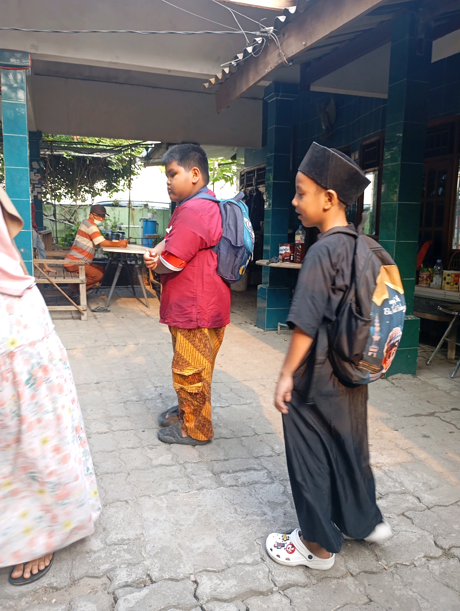 In Babelan, Indonesia, two young boys, likely students, walk side-by-side. The boy on the right wears a black peci (a cap) and a long black garment, carrying a backpack adorned with a cartoon image. The boy on the left sports a red shirt, patterned pants, and a blue backpack. In the background, an adult wearing a striped shirt and mask sits at a table. The scene unfolds on a paved outdoor area with a covered structure overhead, possibly part of a religious or educational institution, with lush greenery visible.