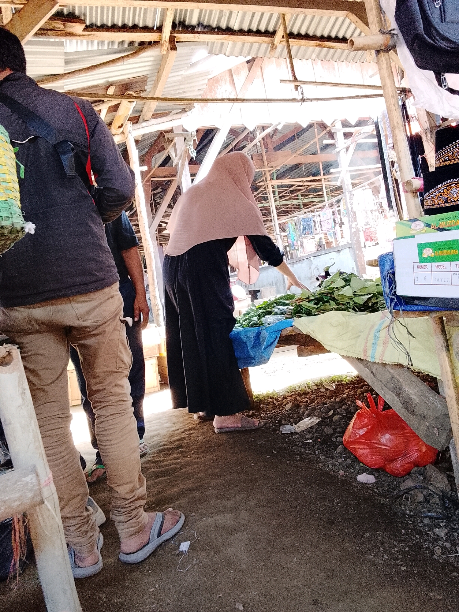 The image depicts an outdoor market scene in Sukarame, Indonesia. In the foreground, a person wearing tan-colored pants and sandals is partially visible from the waist down. To their right, another individual in dark pants and a light pink headscarf is reaching towards a pile of green leaves on a wooden table. The table is covered with a yellow and blue cloth and is set up under a makeshift wooden roof structure. Behind the people, more market stalls and goods are visible, with corrugated metal roofing overhead. A bright red plastic bag is on the ground to the right. The ground is covered in dirt and debris. The lighting suggests it is daytime, possibly sunny, with some areas in shadow.