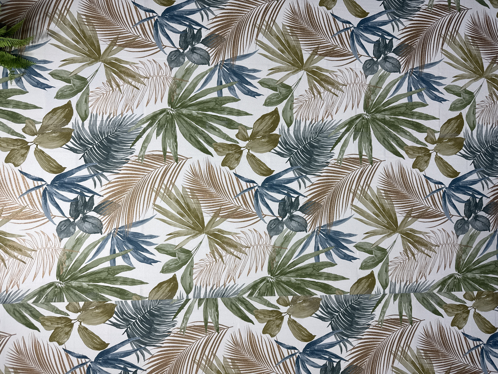 The image is a photograph of a patterned textile, likely wallpaper or fabric, featuring a tropical leaf motif. The design incorporates various types of leaves in shades of green, brown, and blue, with some leaves having a subtle metallic sheen. The leaves are rendered in a watercolor style, creating a soft and artistic effect. The background is a plain white. There are no people or specific landmarks visible in the image. The image does not provide any information to suggest a location in Torrejón de Ardoz, Spain.