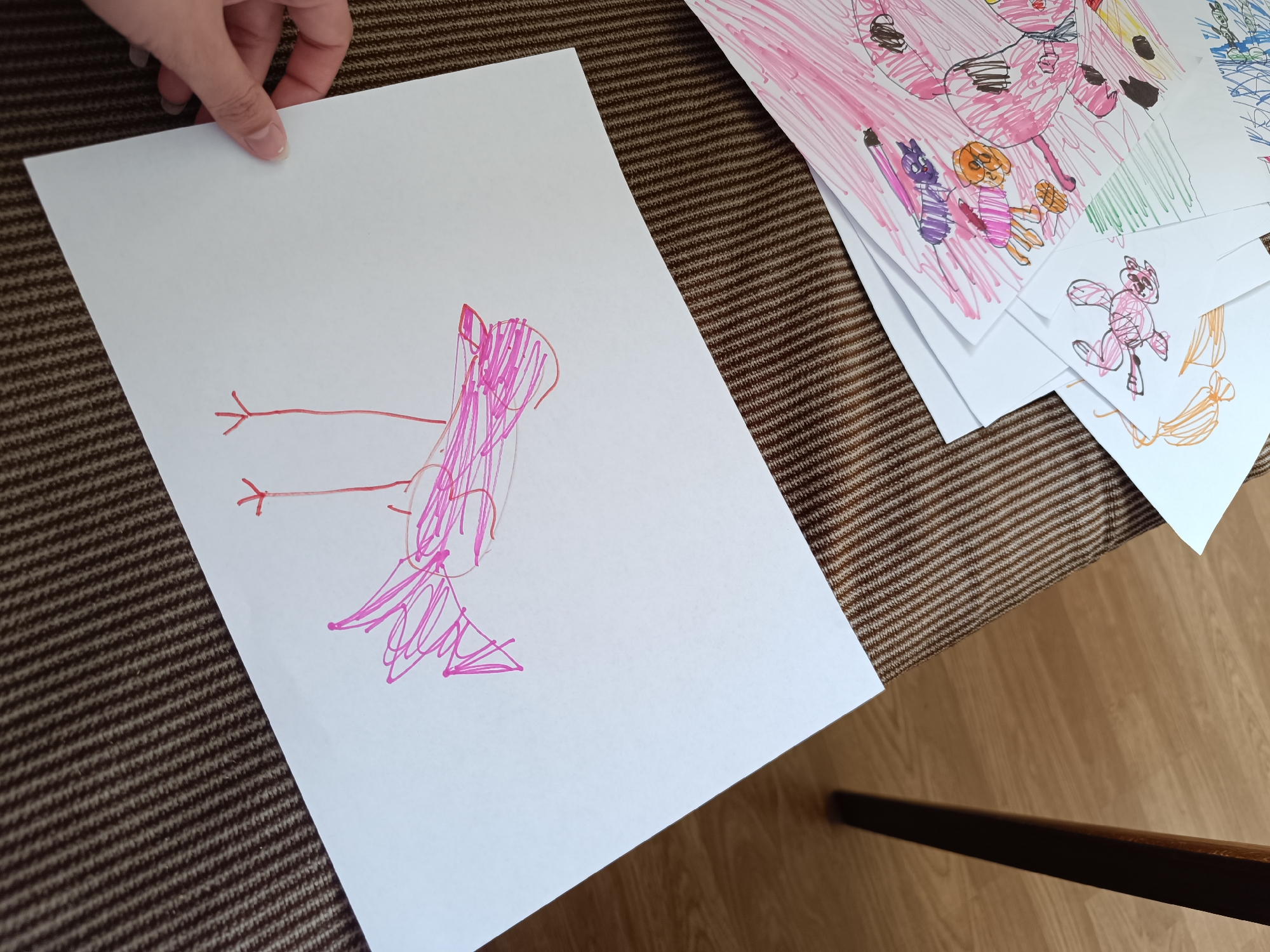 The image shows a brown and tan horizontally striped surface on which several sheets of paper with children's drawings are placed. A hand is holding one sheet of paper with a pink drawing of a bird-like figure with a scribble for its body and simple lines for legs. Other drawings are stacked on top of each other, with visible drawings of a pink cow-like creature, a purple cat-like figure, an orange dog-like figure, and another pink cat-like creature. A portion of a wooden table leg is also visible. The location is Unknown, Unknown.