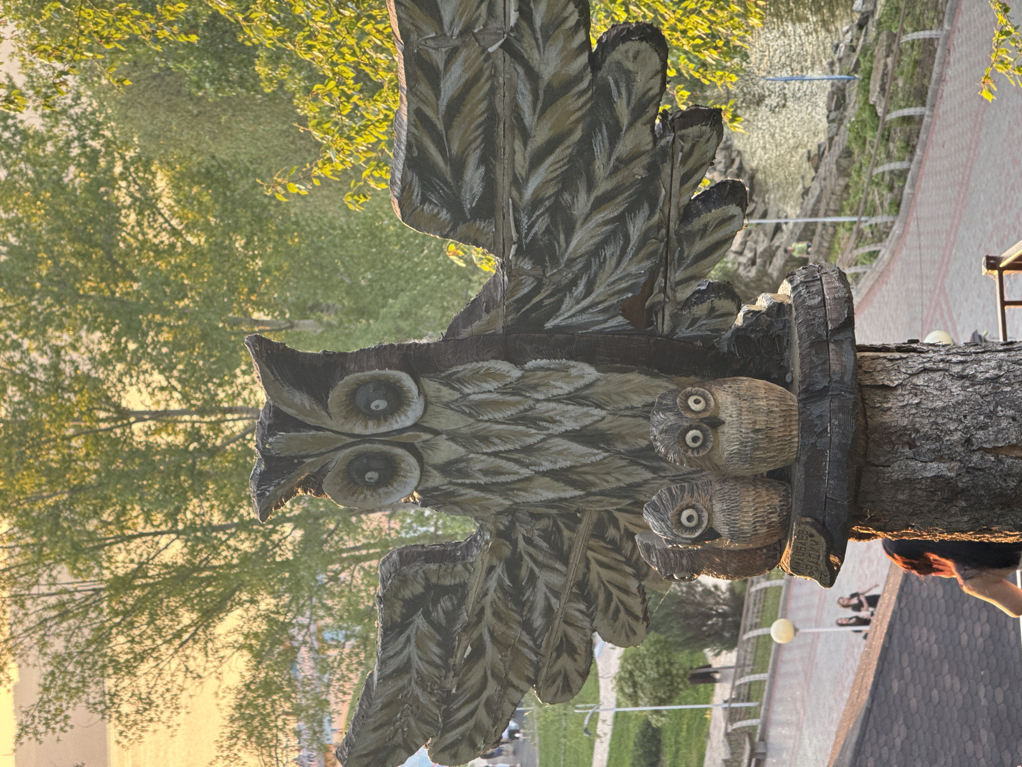 A detailed, carved, wooden sculpture of an owl is the central element of the image. The owl has widespread wings and two smaller, carved owls are positioned on its right side. The sculpture is mounted on a tree trunk base. In the background, blurred scenery depicts trees, a walkway, and some buildings. Indications suggest this is a public area, possibly a park or garden, in Krasny Klyuch, Russia. A person can be seen near the sculpture's base. The overall lighting suggests it is either early morning or late afternoon.