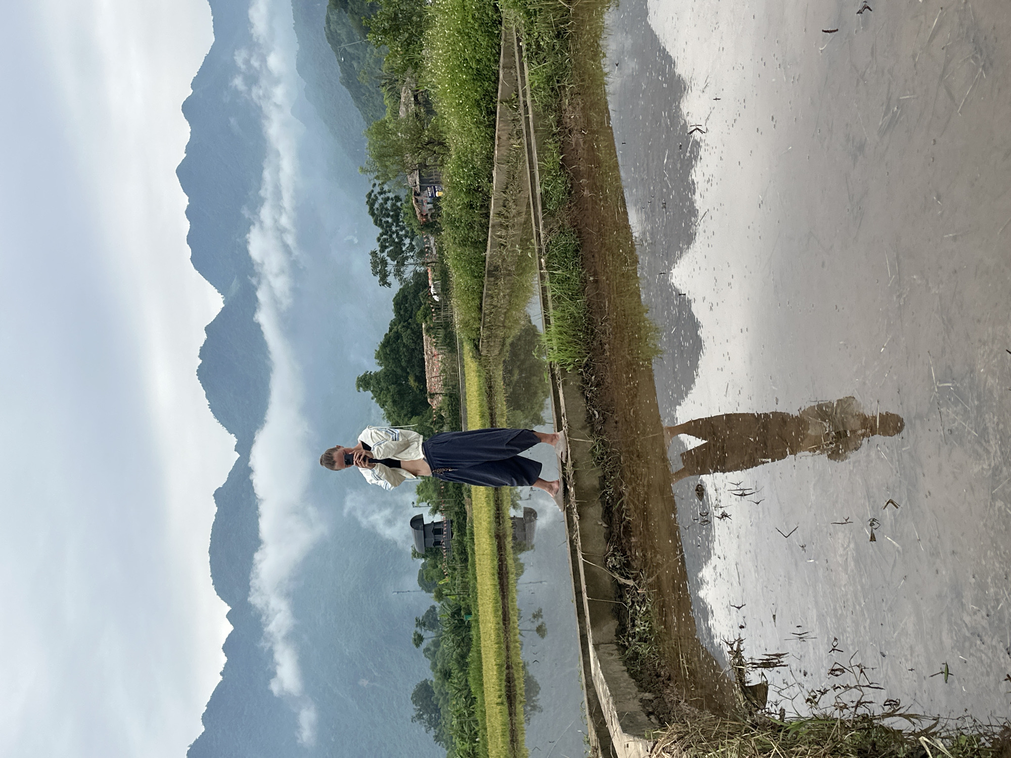 A person stands on a concrete embankment between flooded rice paddies in Mai Chau, Vietnam, holding a mobile phone to take a photo. The person is wearing a white jacket over a black sports bra and loose, dark blue pants. The background features lush green rice fields, trees, and a range of misty mountains under an overcast sky. The water in the paddies reflects the sky, mountains, and the person.
