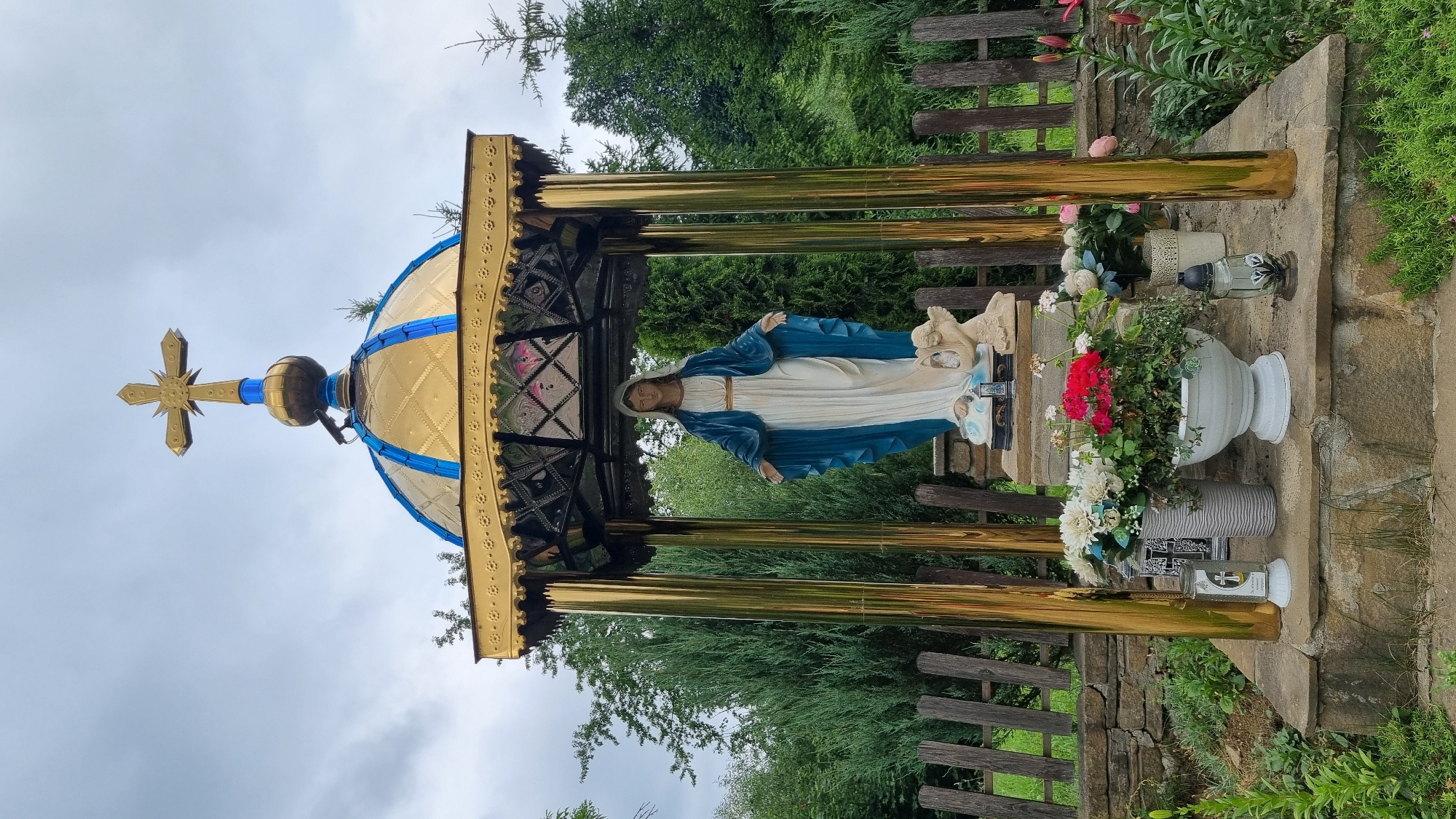 The media file depicts a religious shrine with a statue of the Virgin Mary. The shrine is adorned with a golden dome topped with a cross, and supported by golden pillars. The statue of the Virgin Mary, dressed in a blue mantle and white robe, stands with her arms outstretched. She is accompanied by a smaller figure, possibly representing an angel or child.
The shrine is situated outdoors, amidst lush greenery and trees. A wooden fence runs along the background. In front of the shrine, various offerings are placed, including a large white urn filled with red and white flowers, another vase with white flowers, and a candle holder with a lit candle, featuring Christian symbols. The ground appears to be made of stone. The overall scene suggests a place of worship or remembrance, possibly a roadside shrine or a memorial site.
The lighting suggests it is daytime, and the overcast sky indicates possibly cloudy weather. The location is identified as Vyshka, Ukraine. There are no visible signs of any specific event taking place, other than the presence of religious offerings, which implies ongoing devotion. The mood conveyed is one of reverence and peace. The statue itself shows no visible text, nor does the immediate surroundings, apart from the Christian symbols on the candle holder.