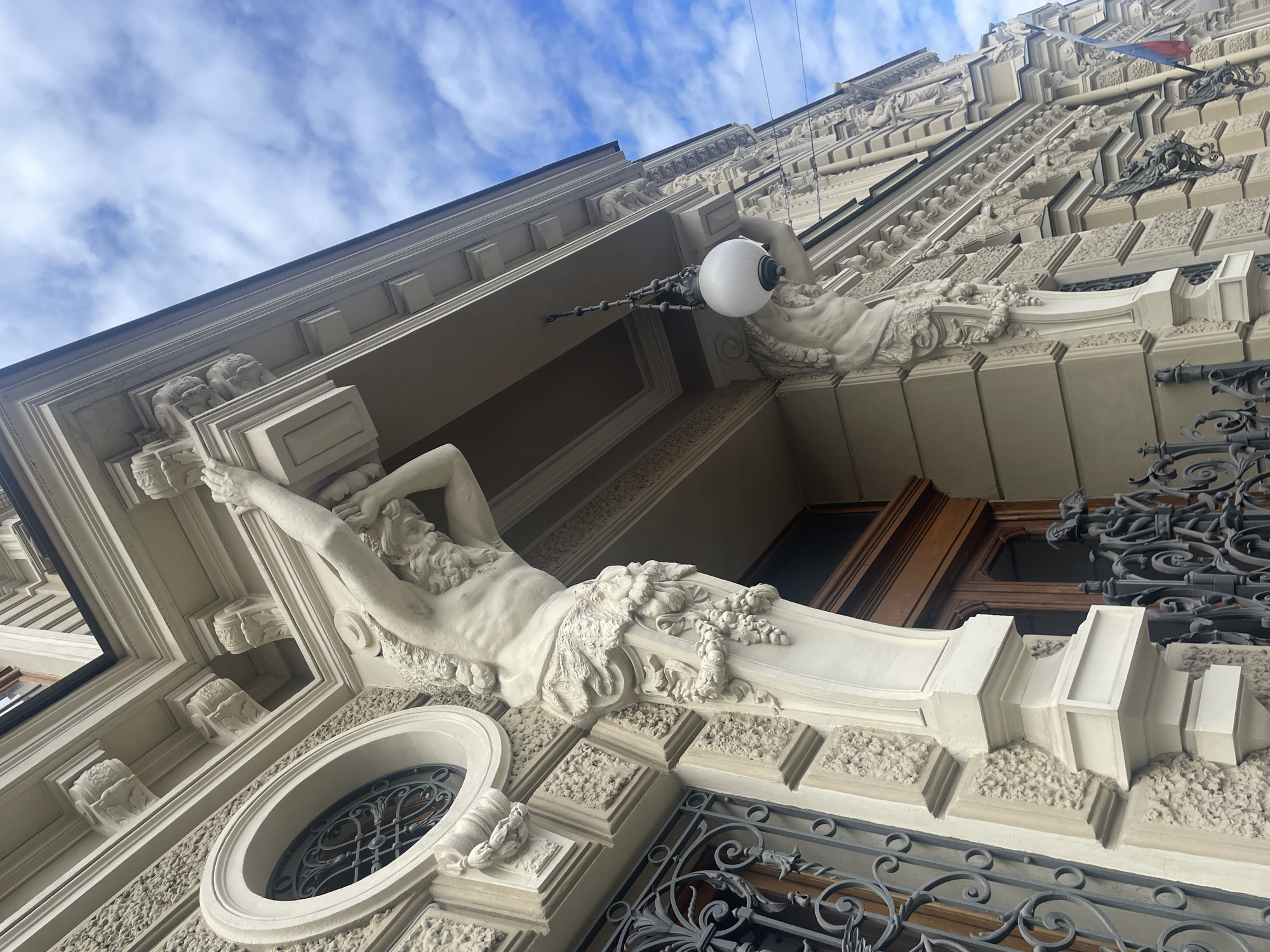 A low-angle, full shot captures the exterior of a grand building in Saint Petersburg, Russia, likely from the 19th century, characterized by its classical architectural style. The facade is adorned with ornate sculptures of Atlas figures, supporting the building's upper story. These figures are depicted with muscular torsos, flowing beards, and are partially draped in foliage. The building's stonework features intricate detailing, including carved moldings, decorative grilles over windows, and a large, round window with a wrought-iron grille. A spherical lamp hangs from a chain beneath an overhang, also supported by a sculpted figure. The sky above is a mix of blue with scattered white clouds. A Russian flag is visible in the distance on the right side of the building. The overall impression is one of historical grandeur and architectural artistry.
