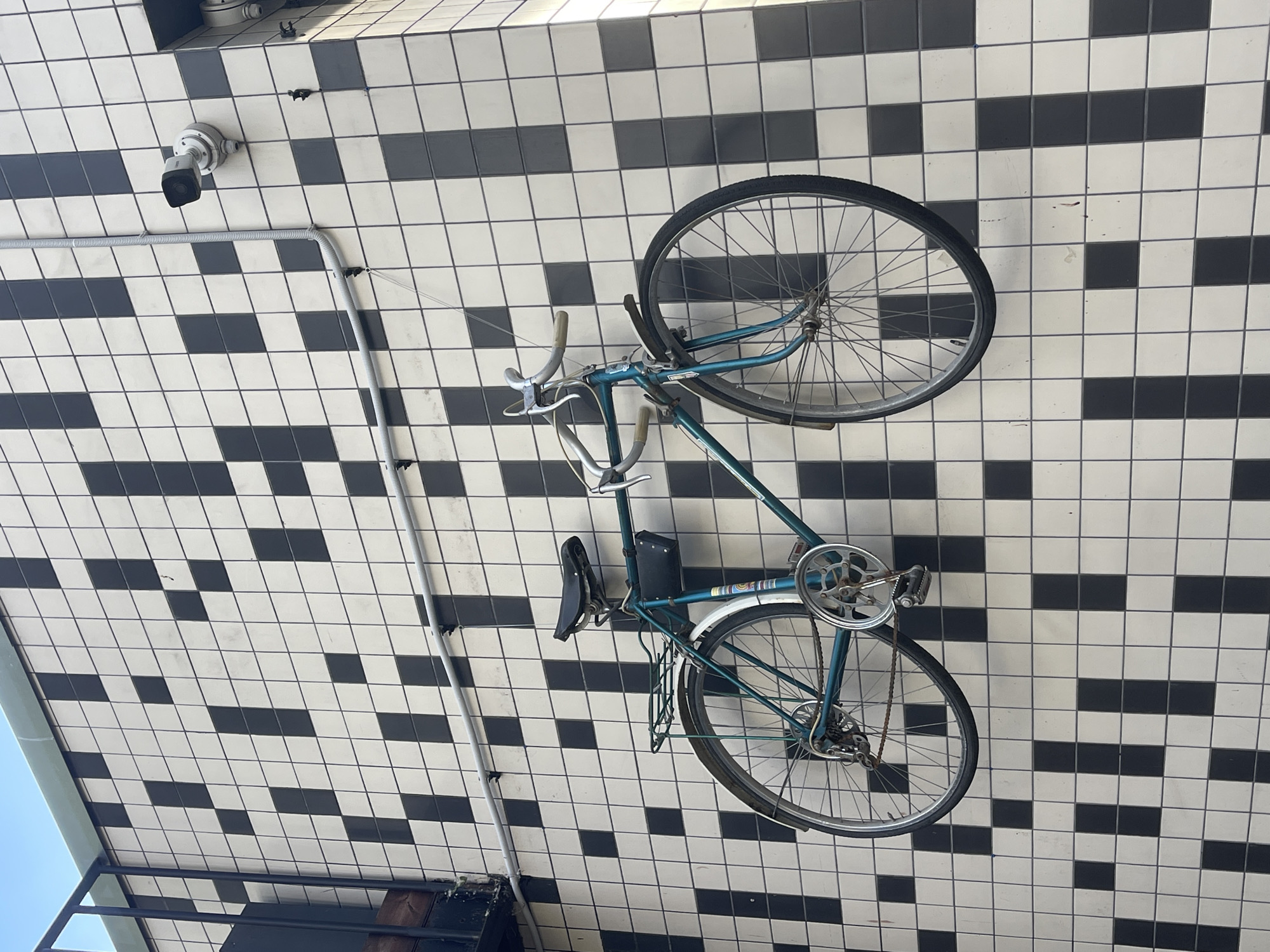 The image shows a vintage teal bicycle suspended from a tiled ceiling. The ceiling is adorned with a checkered pattern of black and white square tiles, creating a visually striking background. The bicycle is the primary object of focus, with its frame, wheels, handlebars, and pedals clearly visible. A security camera is mounted on the tiled ceiling to the upper left of the bicycle. The lighting suggests it is daytime, possibly indoors or under an overhang, given the even illumination and lack of strong shadows. The overall scene appears to be in an urban or commercial setting, given the presence of the security camera and the tiled architecture. There are no people visible in the image. The bicycle is stationary, implying it is displayed or stored in this manner. There is no visible text in the image.